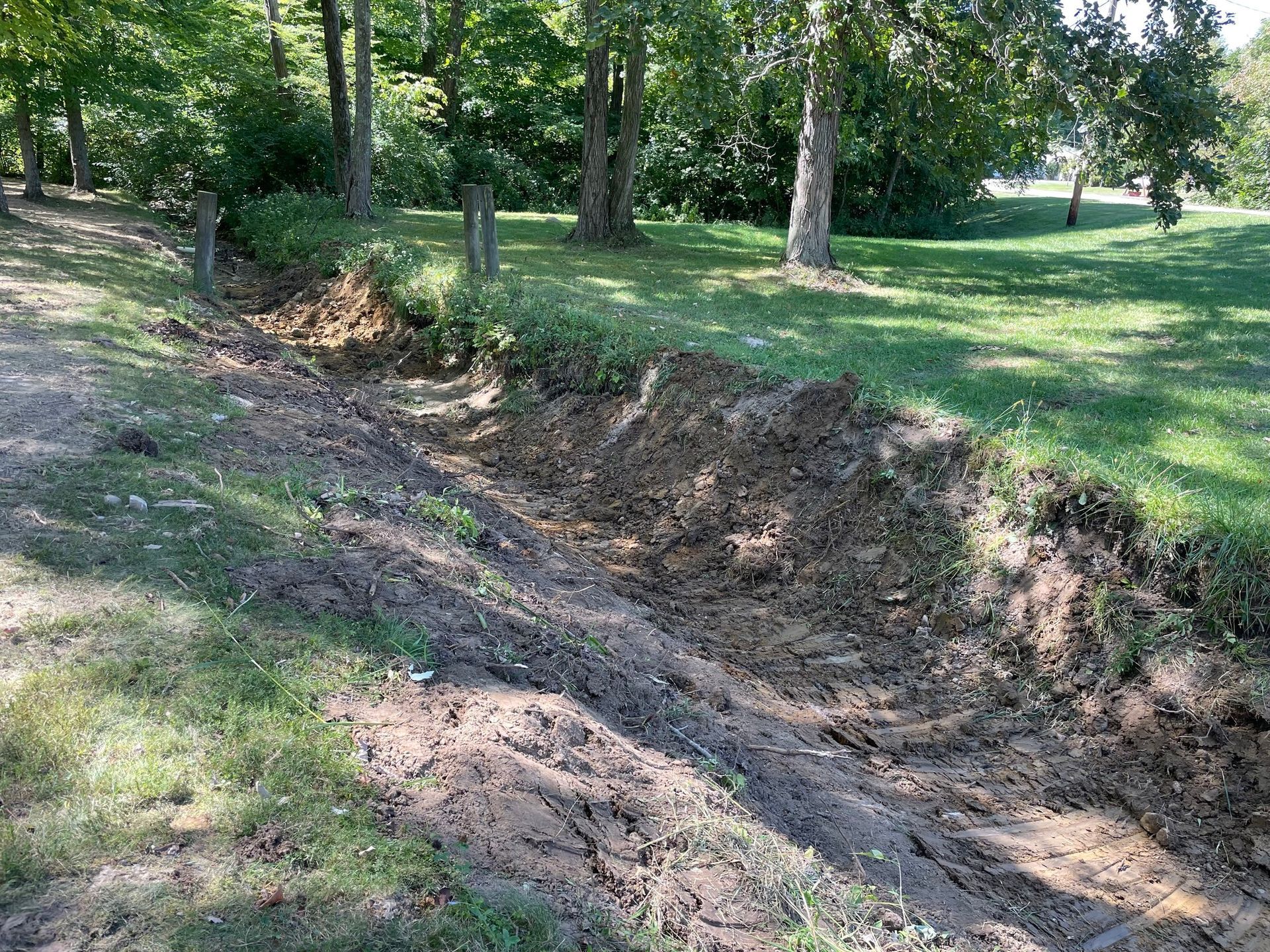 Dirt drainage ditch along a grassy, tree-lined park edge on a sunny day