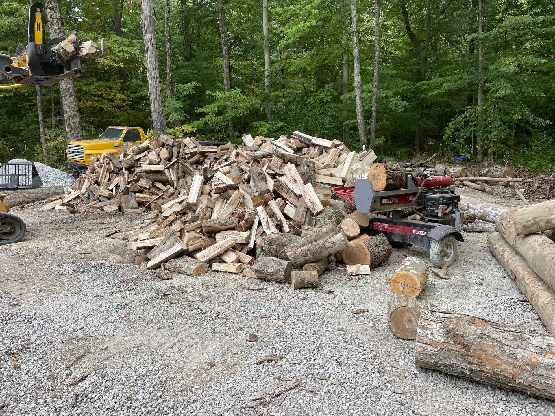 Pile of chopped firewood and logs at a gravel clearing beside a wooded area, with a log splitter nearby.