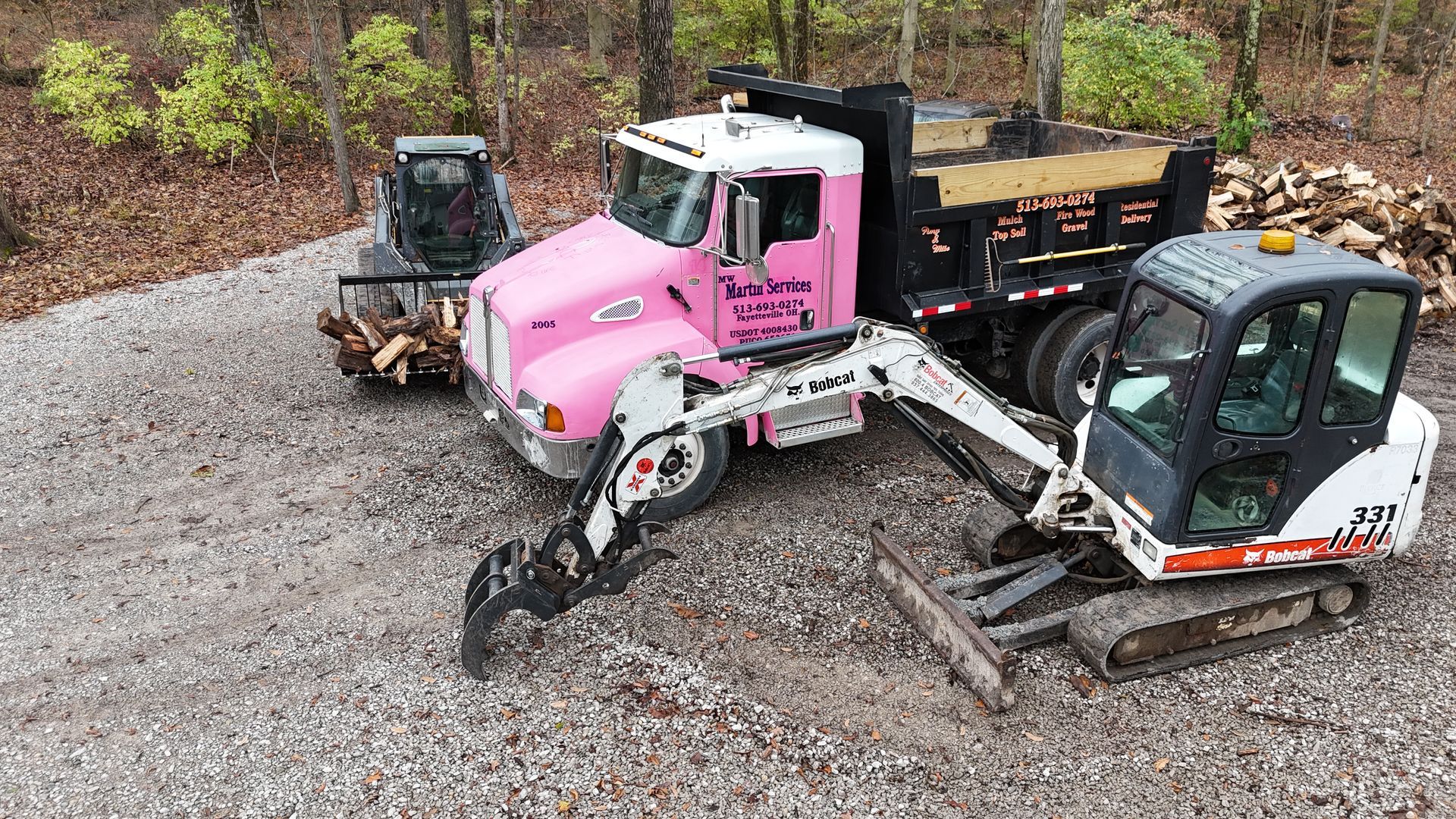 Pink and black construction vehicles and a small excavator on a gravel lot beside a wooded area