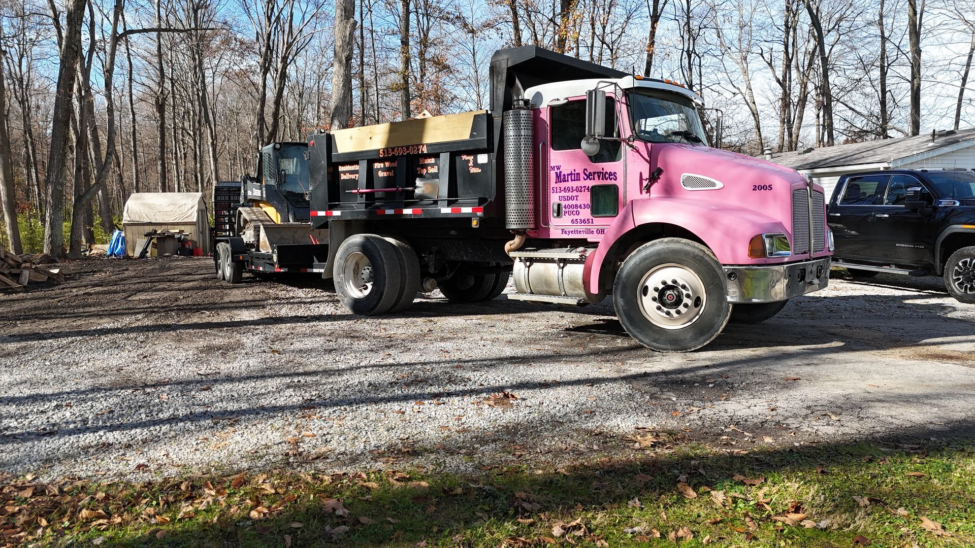 Pink dump truck parked on gravel driveway beside a black SUV, with bare trees in the background