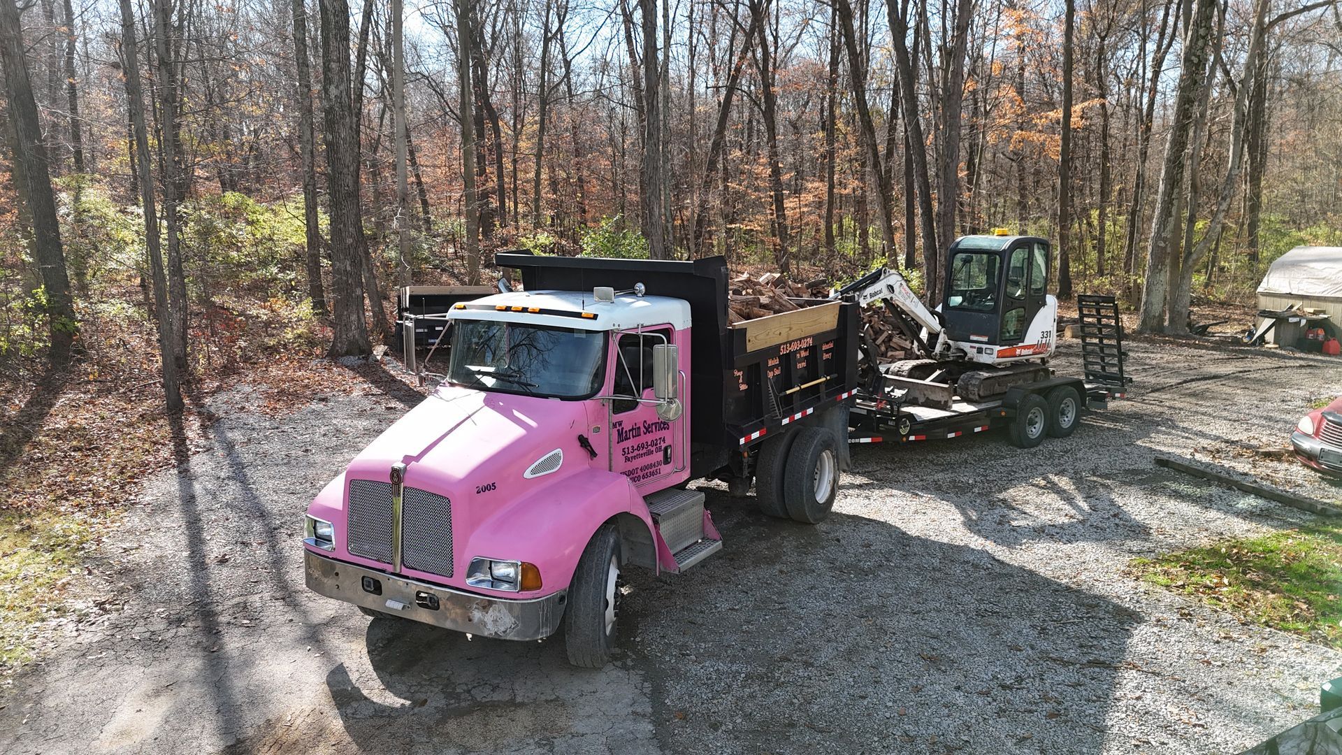 Pink dump truck hauling a skid steer on a gravel driveway in a wooded yard