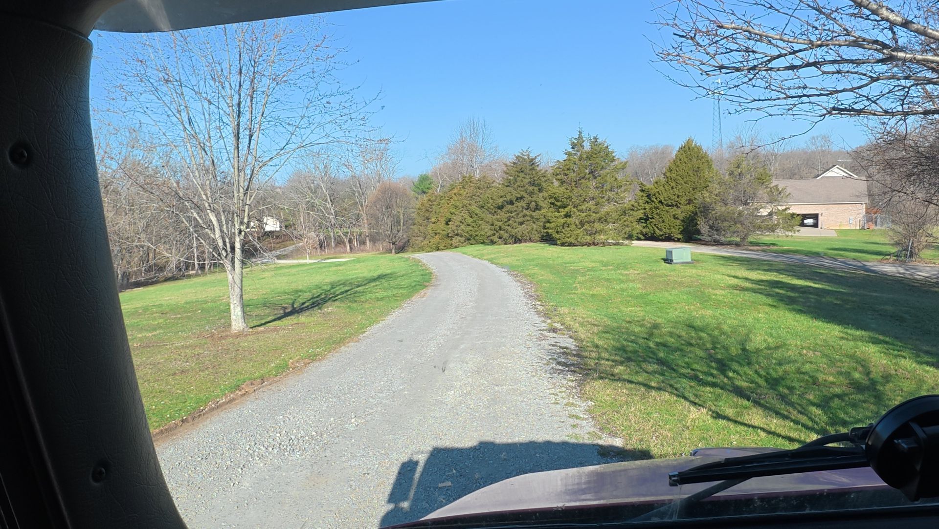Gravel driveway curving through a green yard with trees and a house on the right, viewed from a vehicle.