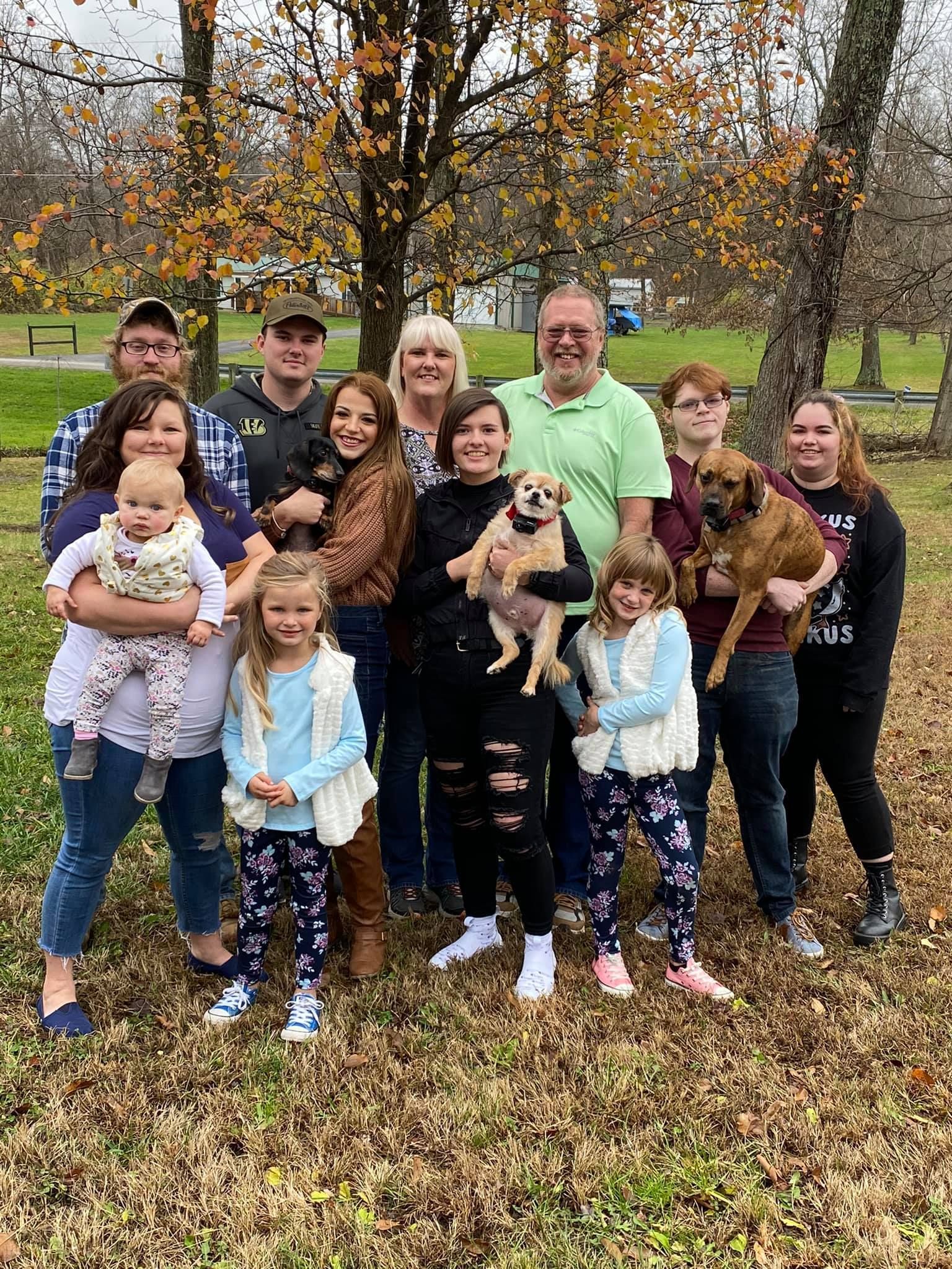 Large family posing outdoors in autumn, with children, dogs, and a baby in front of trees.
