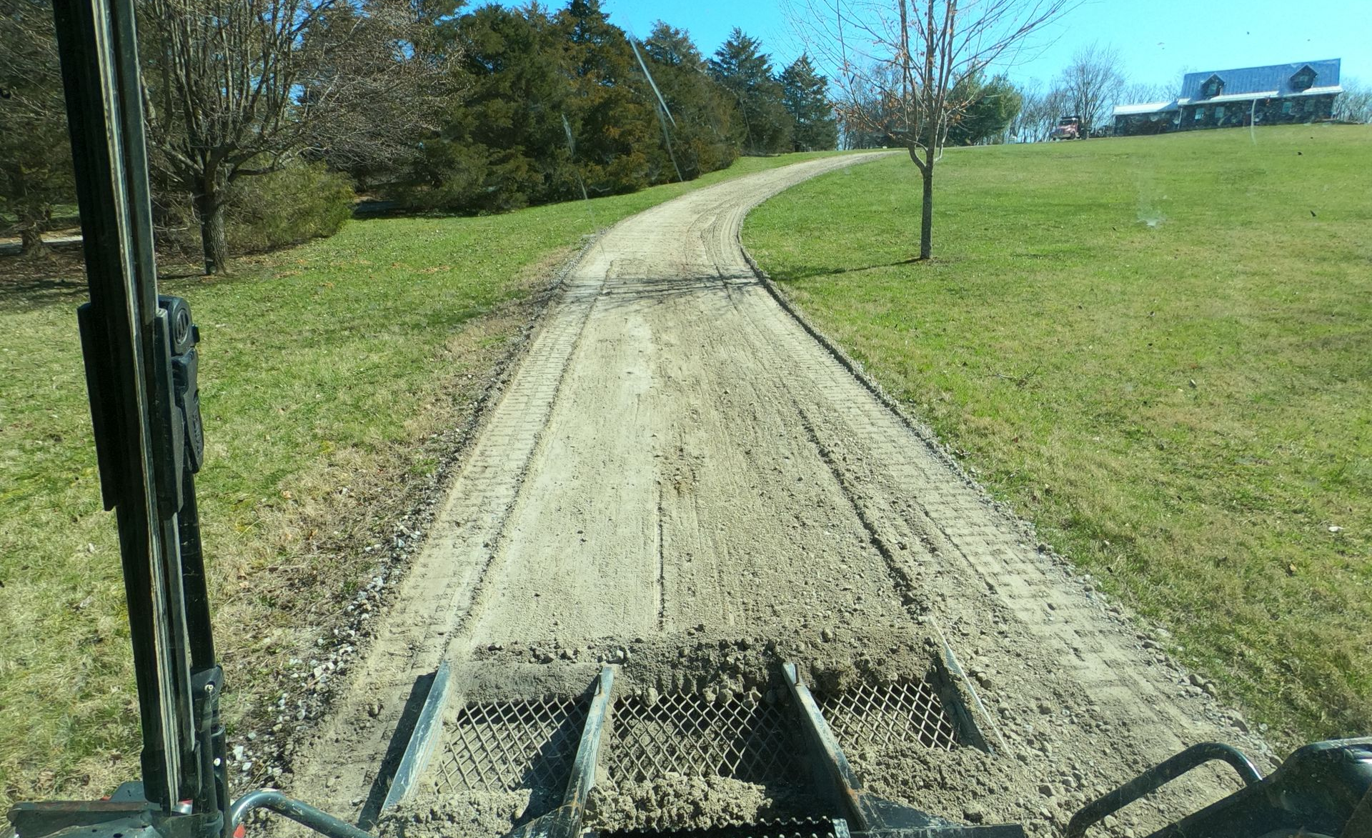 Gravel path curving through a sunny grassy field, viewed from a vehicle dashboard.