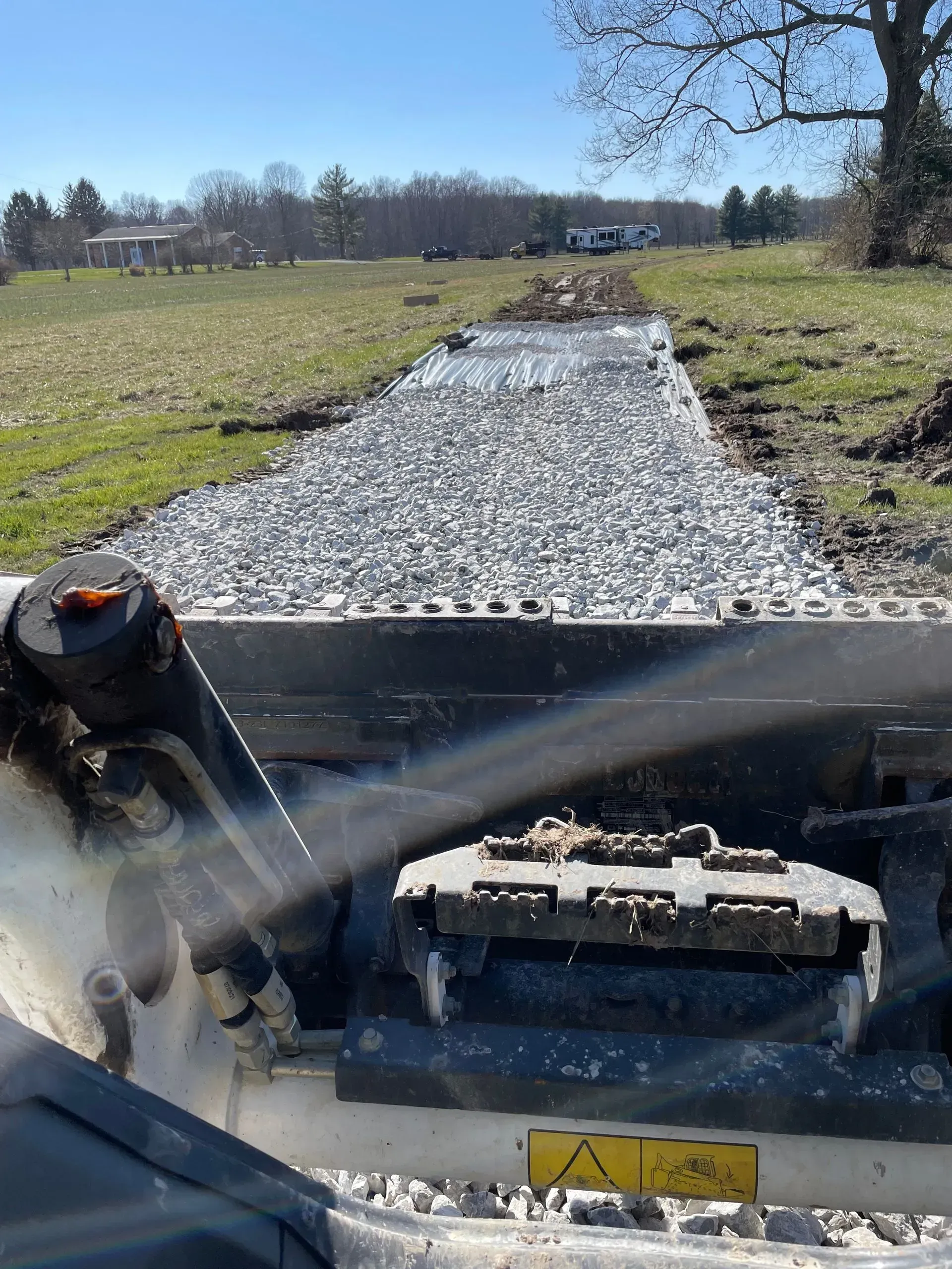 Construction vehicle spreading gravel on a rural path beside open fields under a clear blue sky