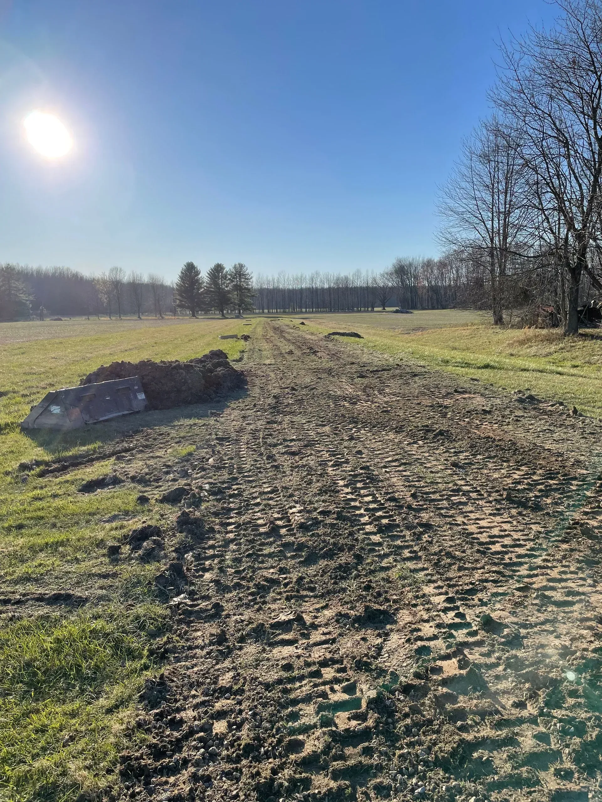 Dirt path through a sunny grassy field with trees and a pile of soil on the left.