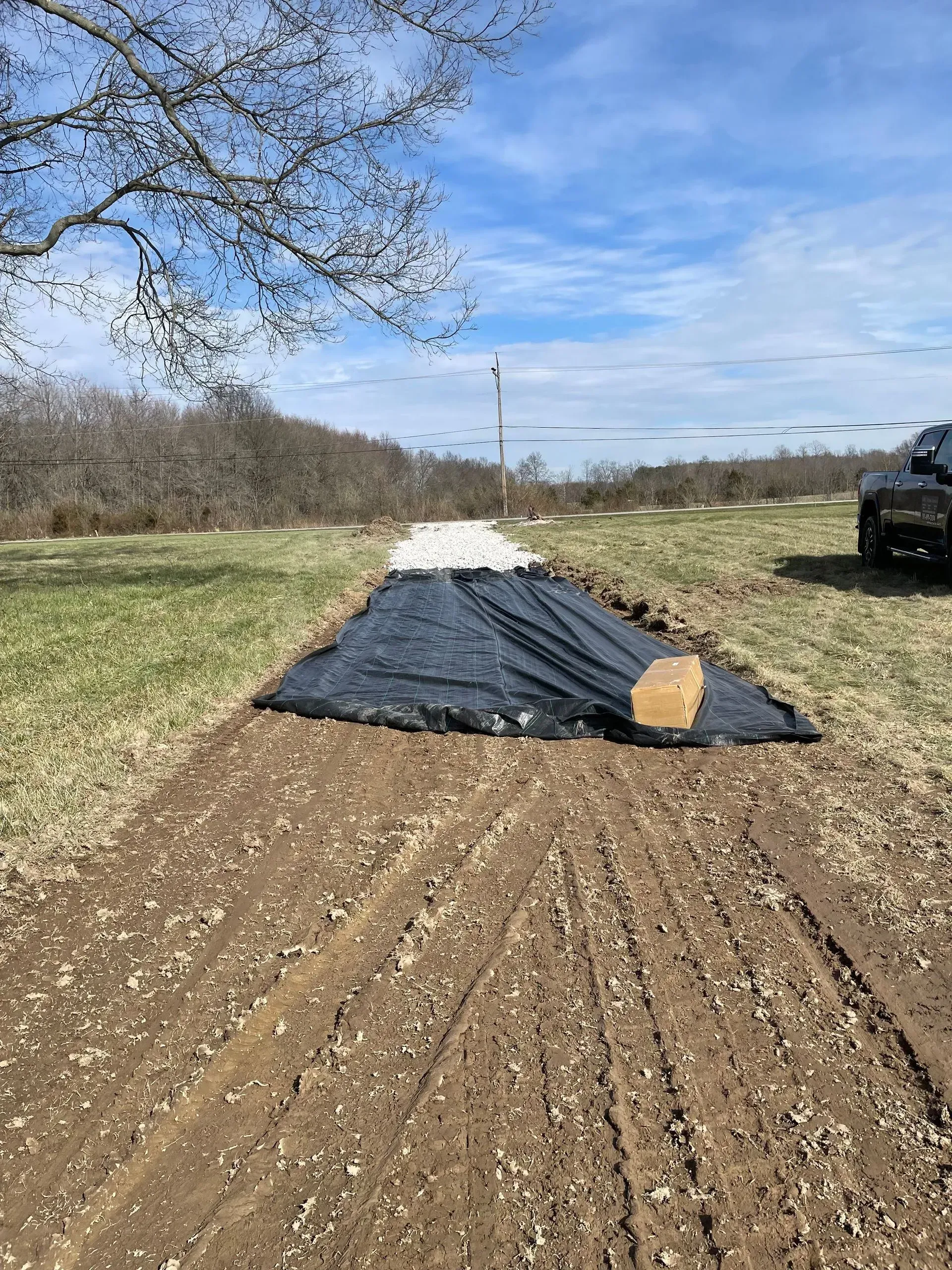 Black tarp spread on a dirt road beside a grassy field under a blue sky, with a vehicle at right.