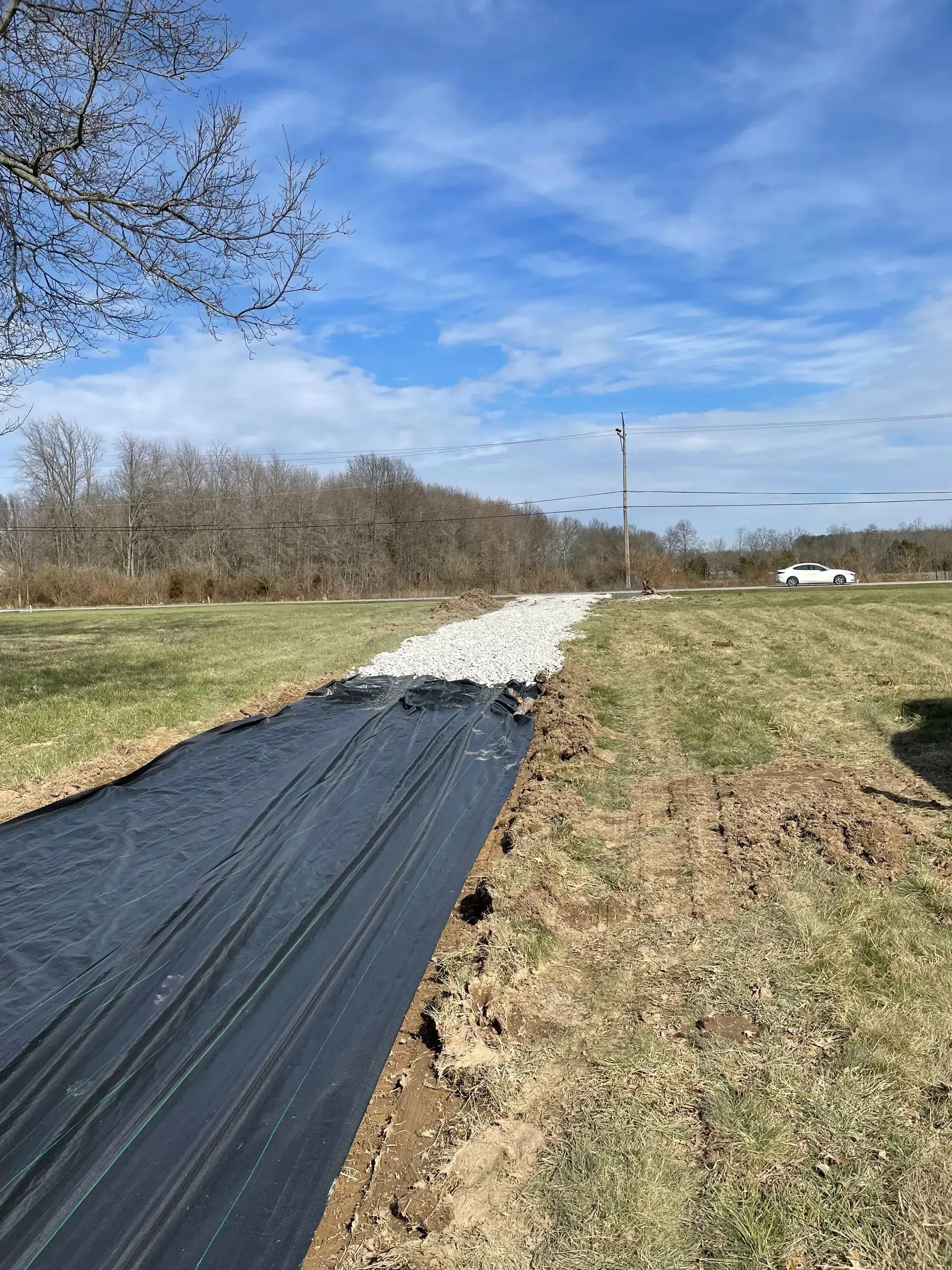 Black tarp-lined path through grassy field under blue sky, with gravel ahead and a distant utility pole