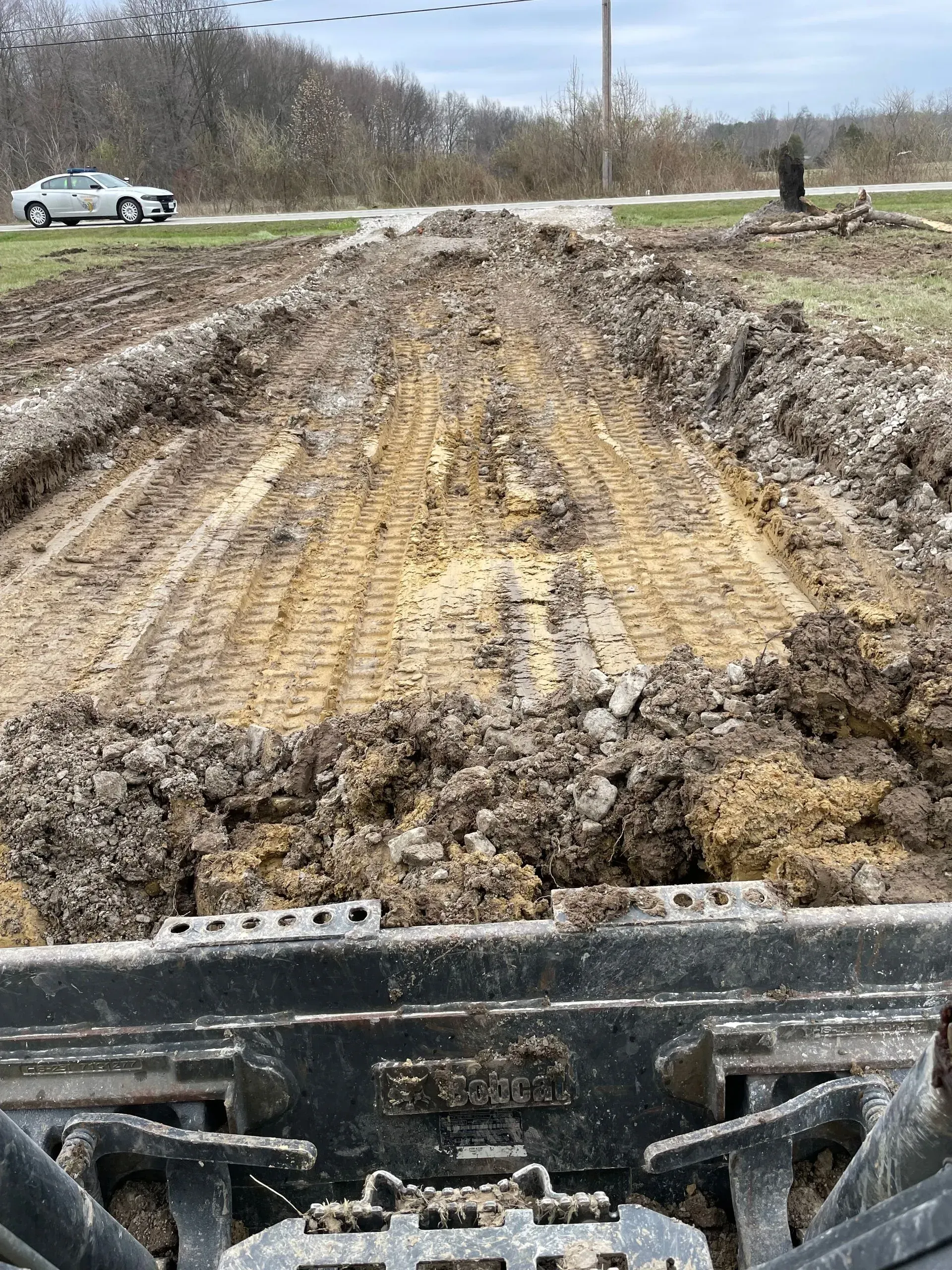 Muddy construction road viewed from a vehicle, with tire tracks and a car on the left roadside.