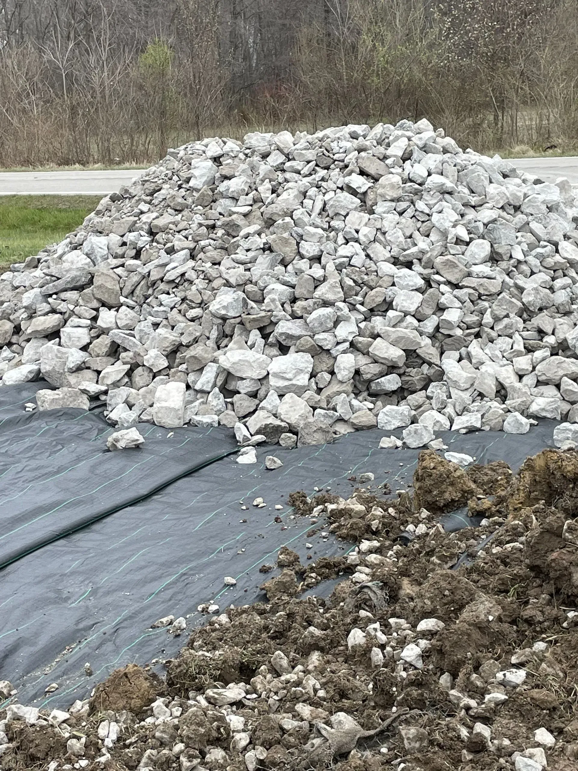Large pile of gray rocks on black landscape fabric beside muddy ground outdoors