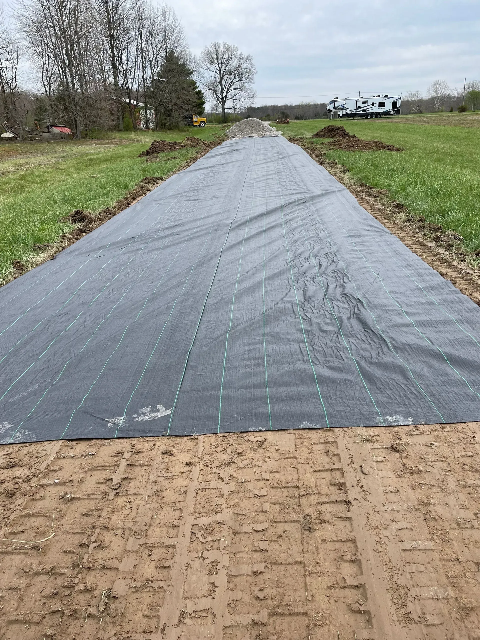 Black landscape fabric laid over a long gravel path in a grassy field under cloudy skies