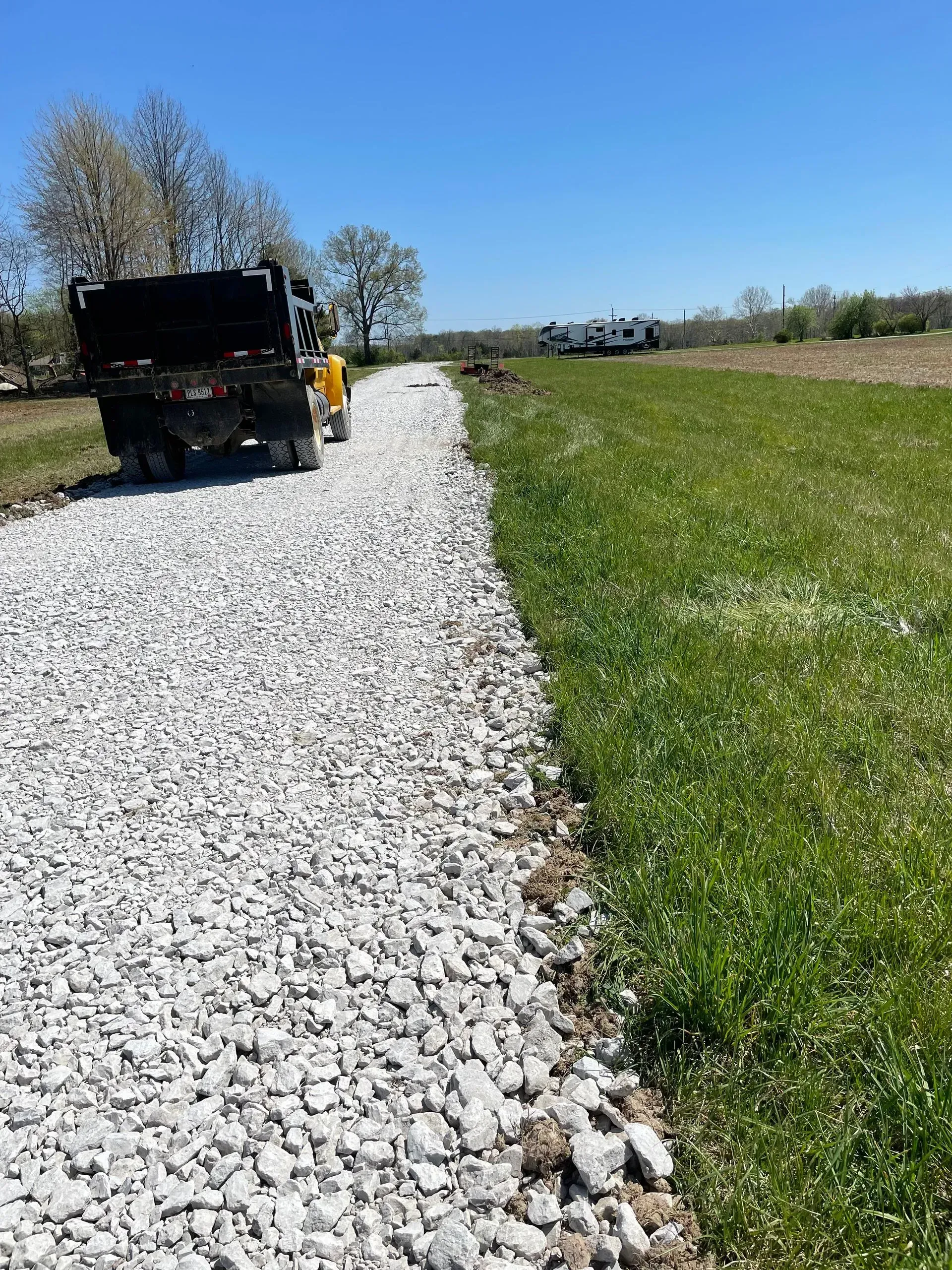 Gravel road under construction beside a green field, with a dump truck and worker on a sunny day