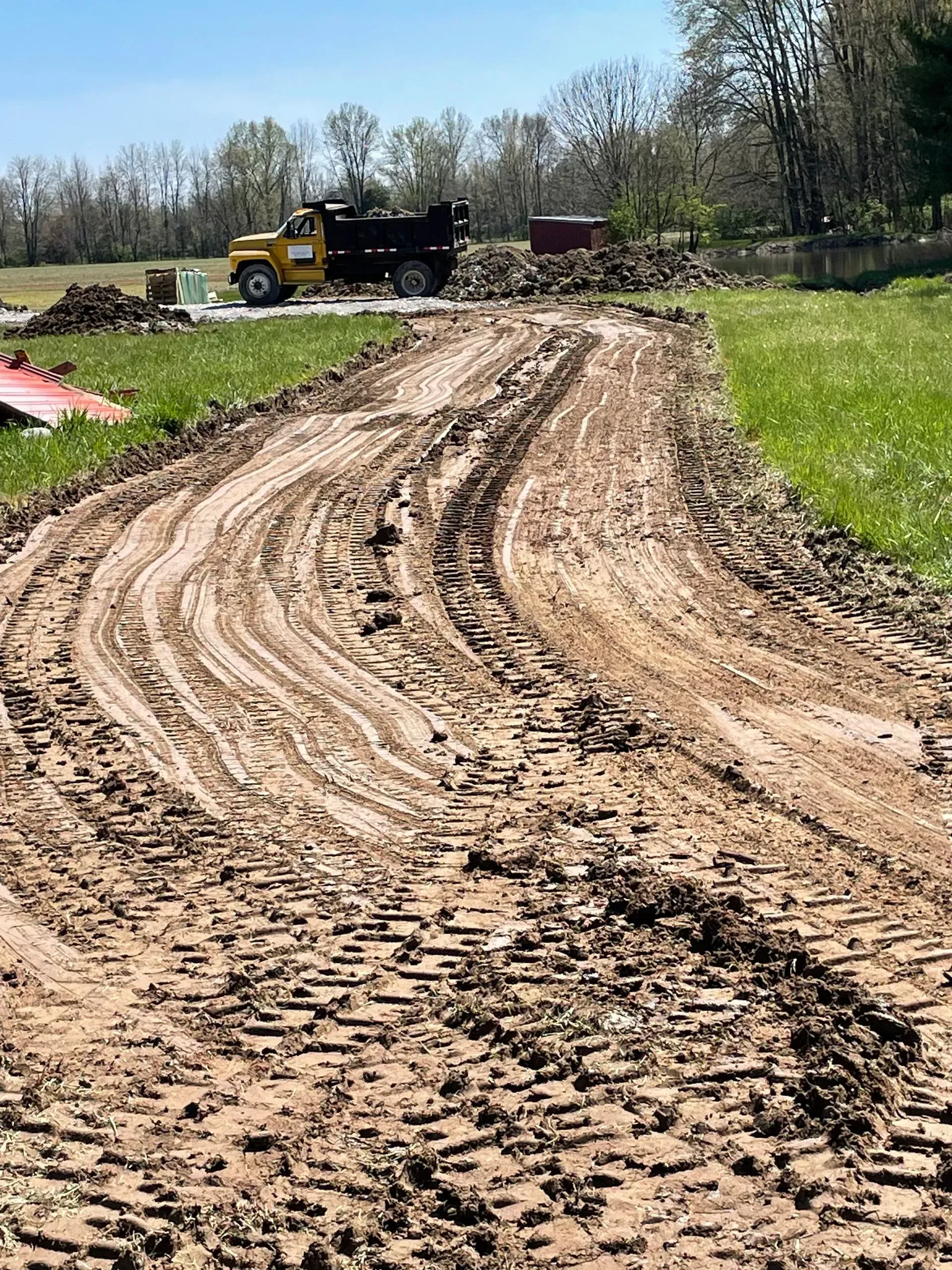 Muddy dirt road with tire tracks beside a yellow dump truck in a rural field.