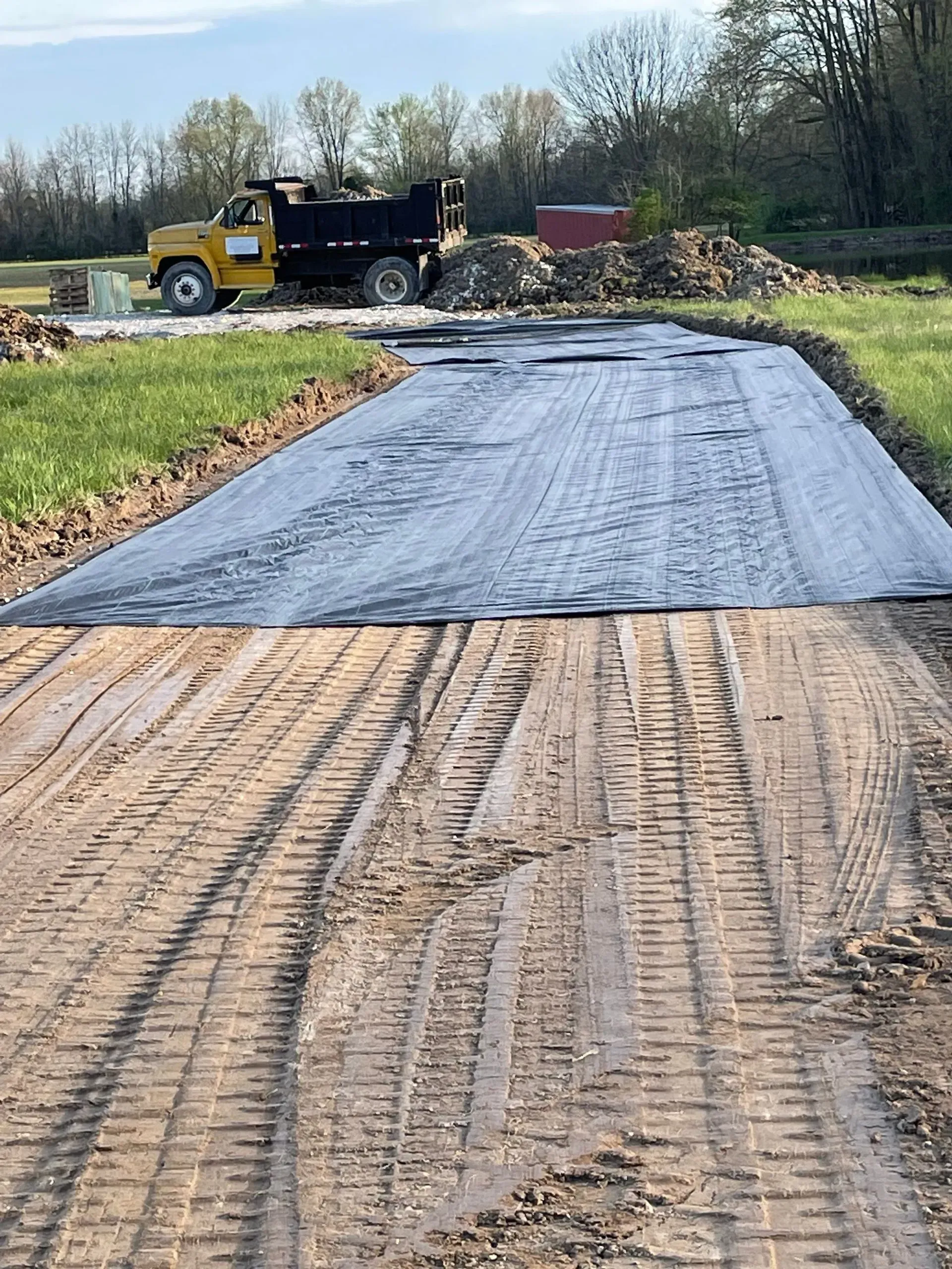 Black geotextile laid on a dirt road at a construction site with a dump truck and trees in the background