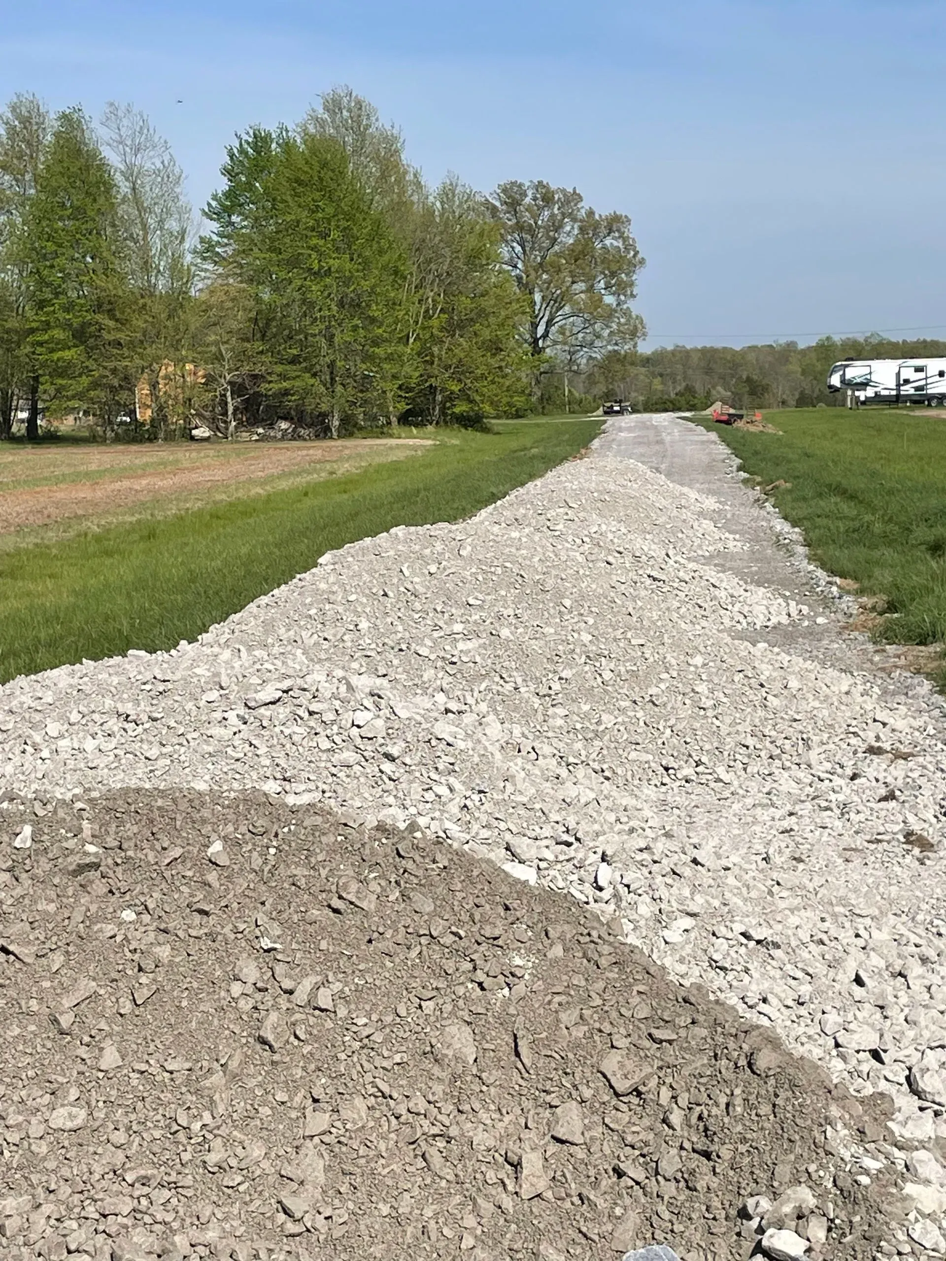 Gravel path under construction beside a grassy field with trees and parked vehicles in the distance
