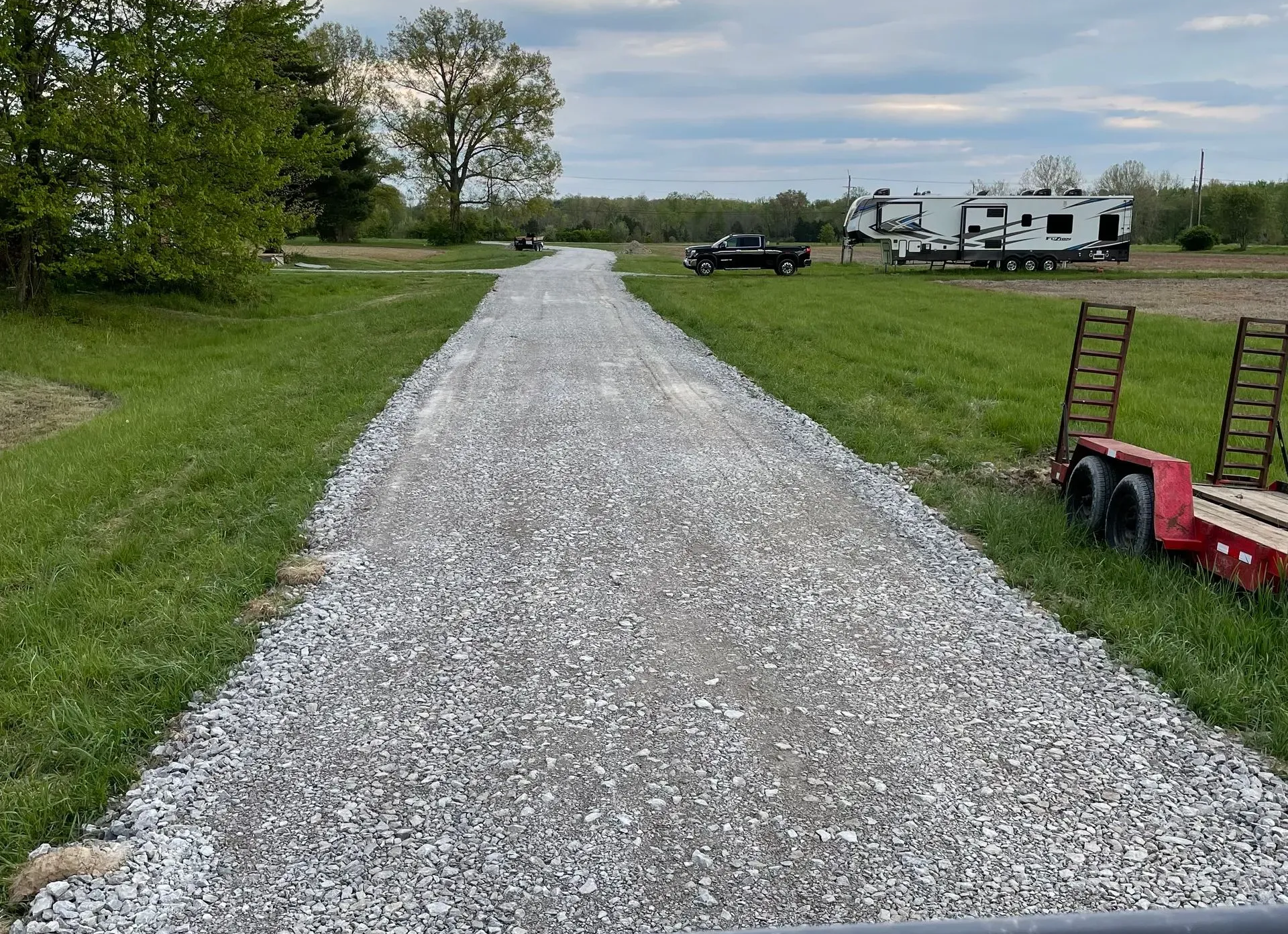 Gravel driveway leading to RVs in a grassy campground, with a red utility trailer on the right.