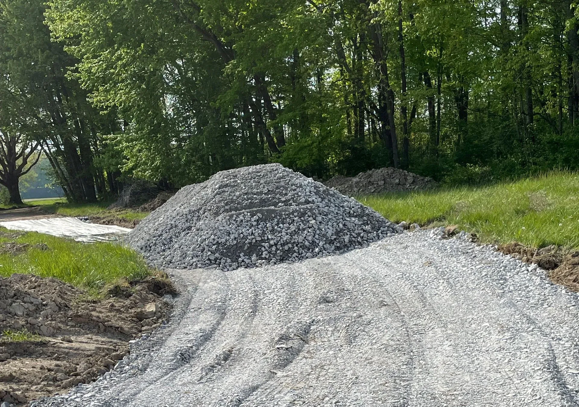 Gravel driveway under construction with a large pile of crushed stone beside a wooded roadside