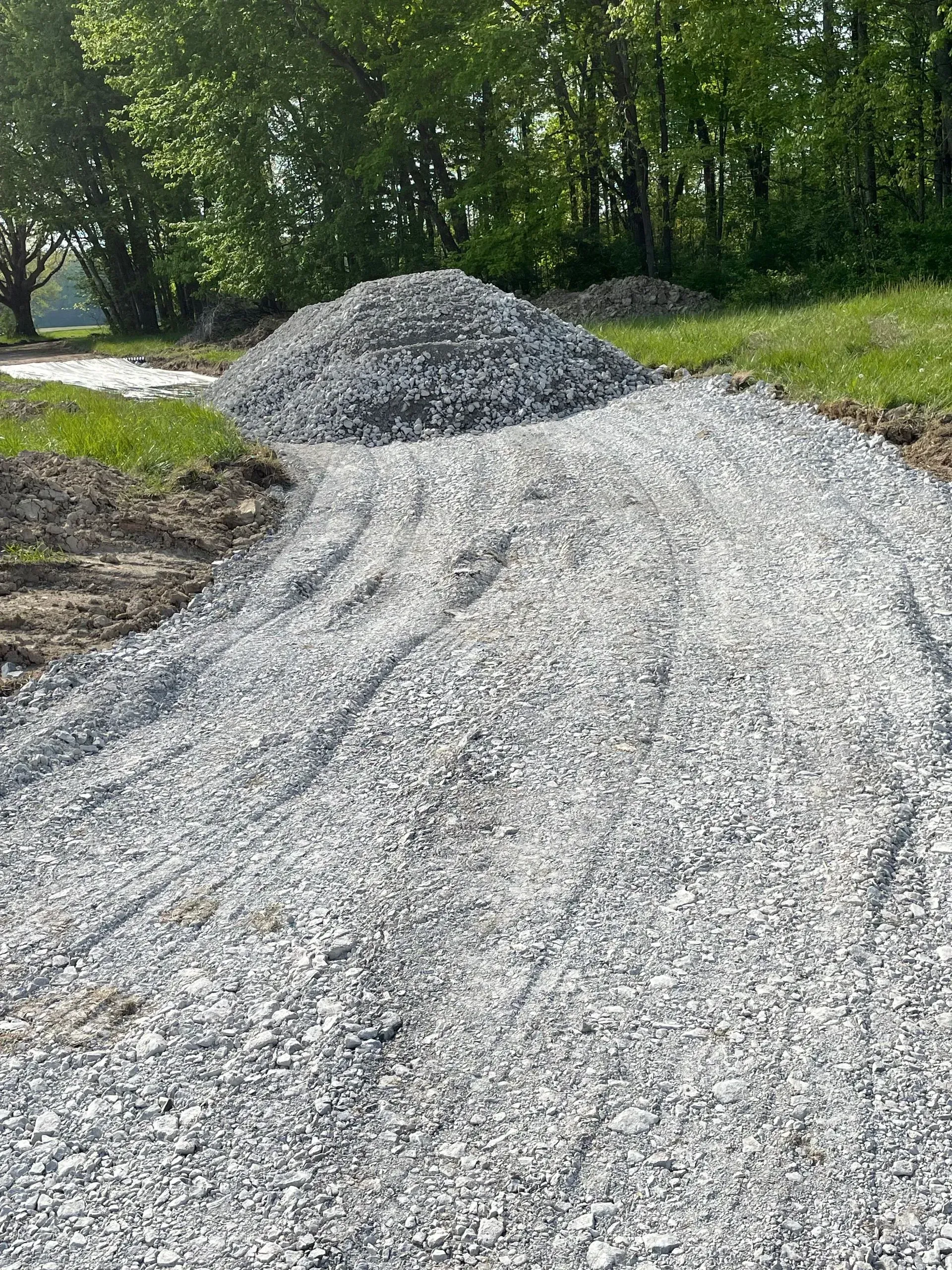 Gravel pile beside a rough gravel driveway in a wooded area on a sunny day