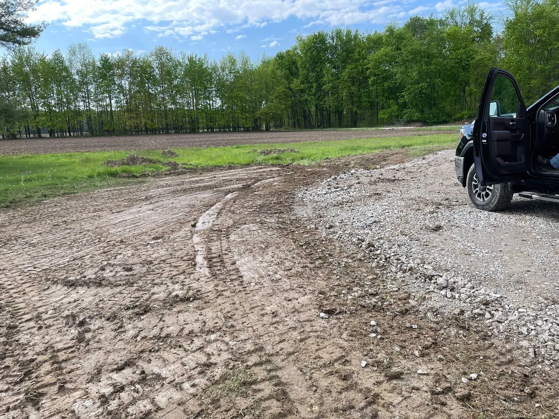 Muddy dirt road beside a green field and trees, with an open black vehicle at right.