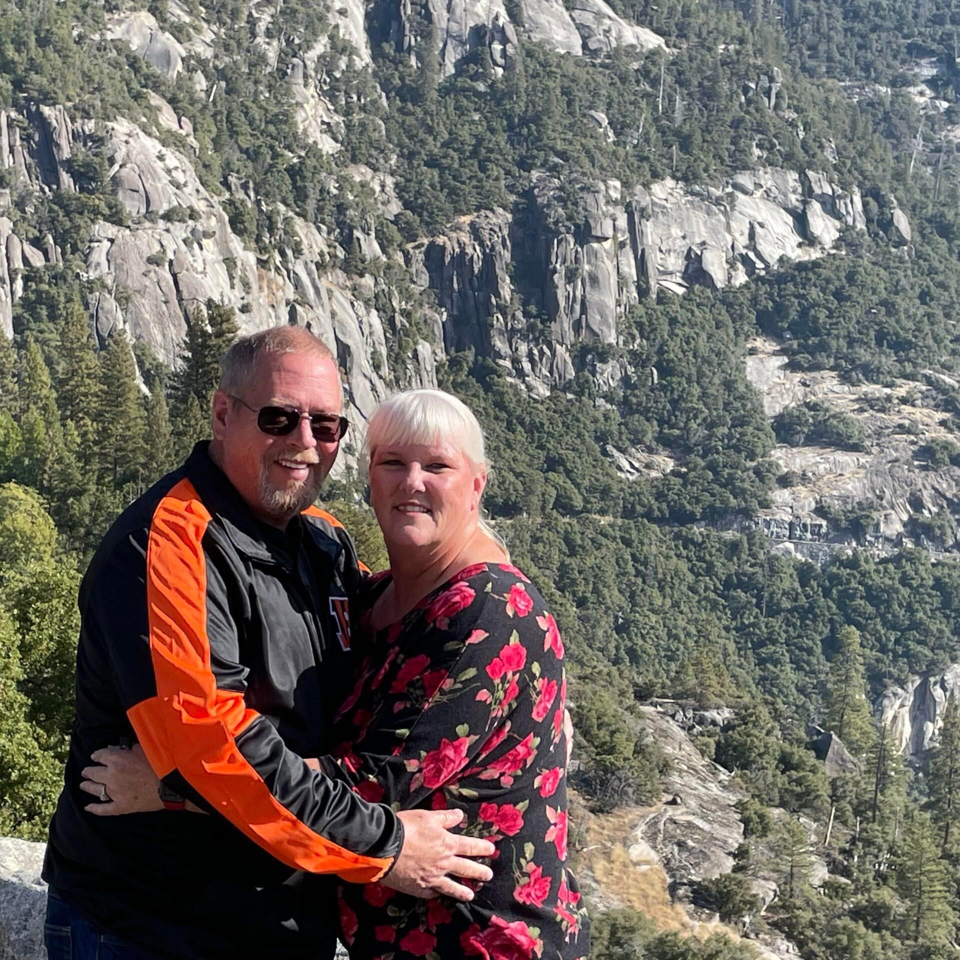 Smiling couple posing on a mountain overlook with rocky cliffs and pine trees in the background