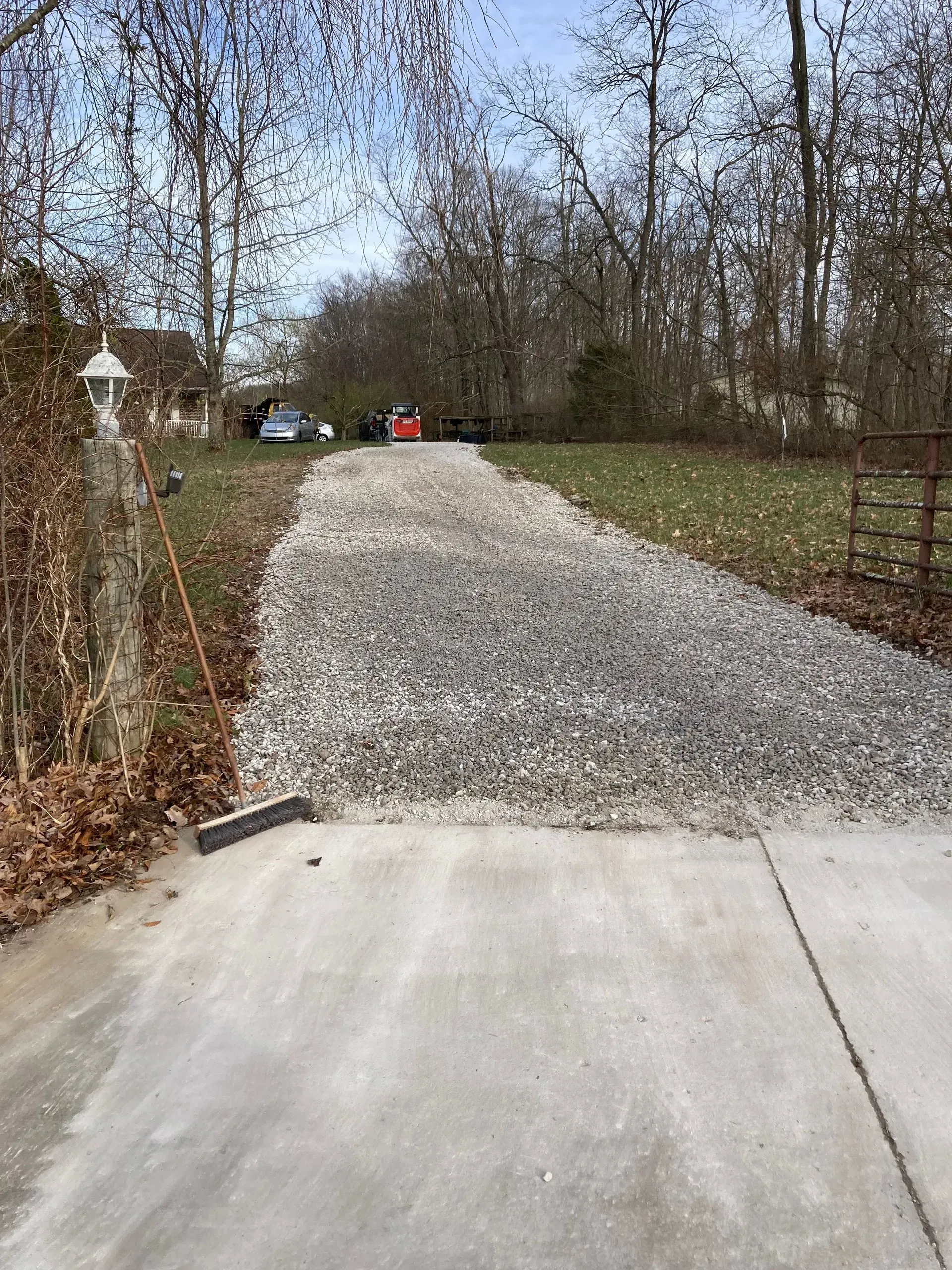 Gravel driveway with concrete apron, trees, lamp post, and fence along a wooded yard