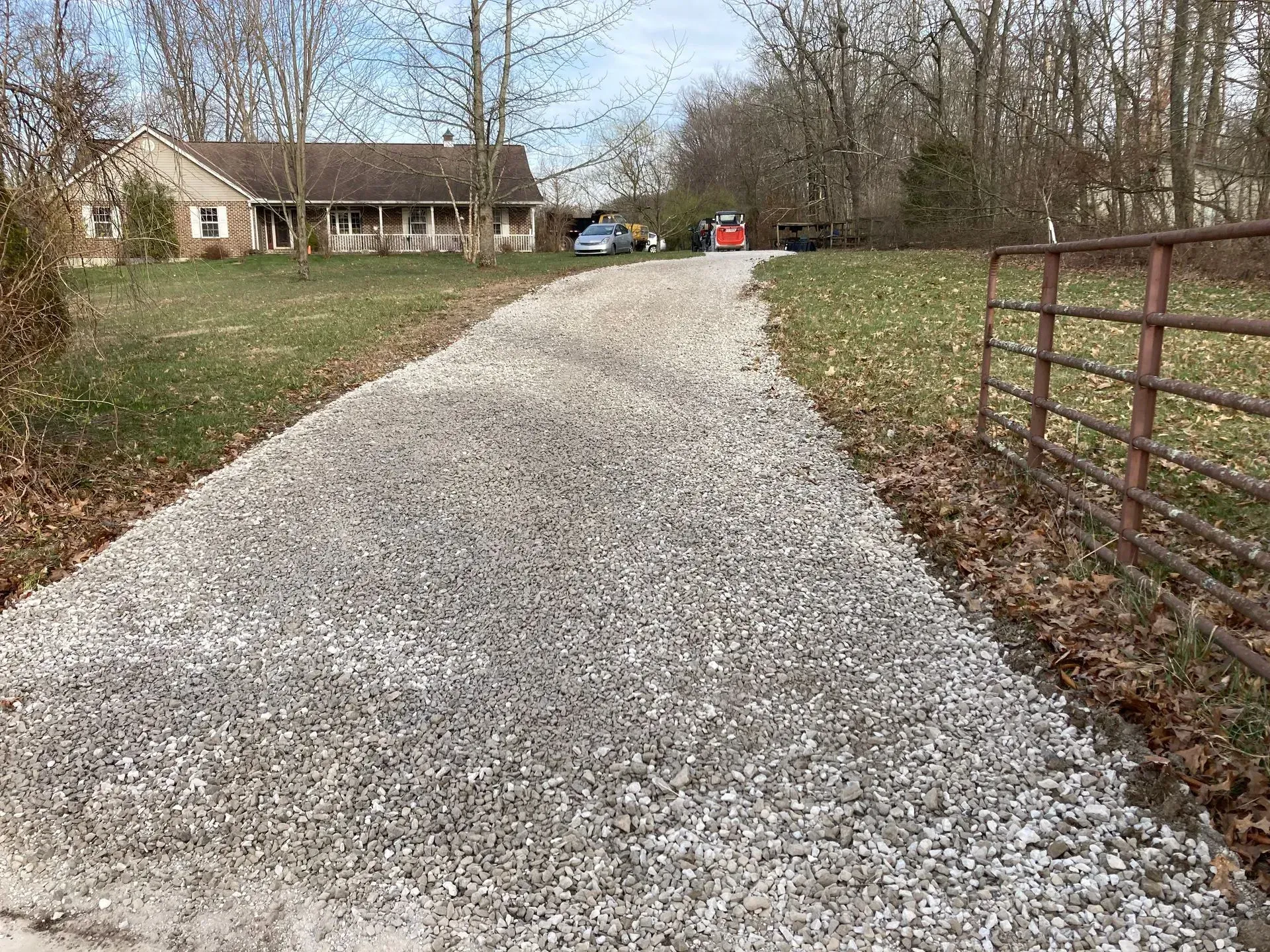 Gravel driveway leading to a house, with leafless trees and a red gate on the right.