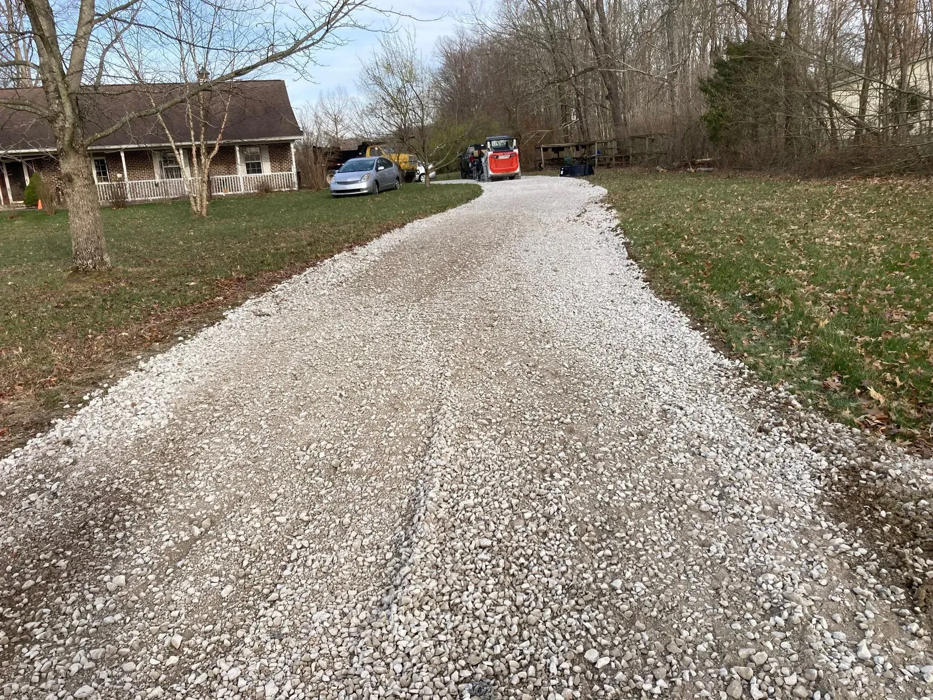 Gravel driveway leading to a rural house, with parked vehicles and leafless trees on a cloudy day