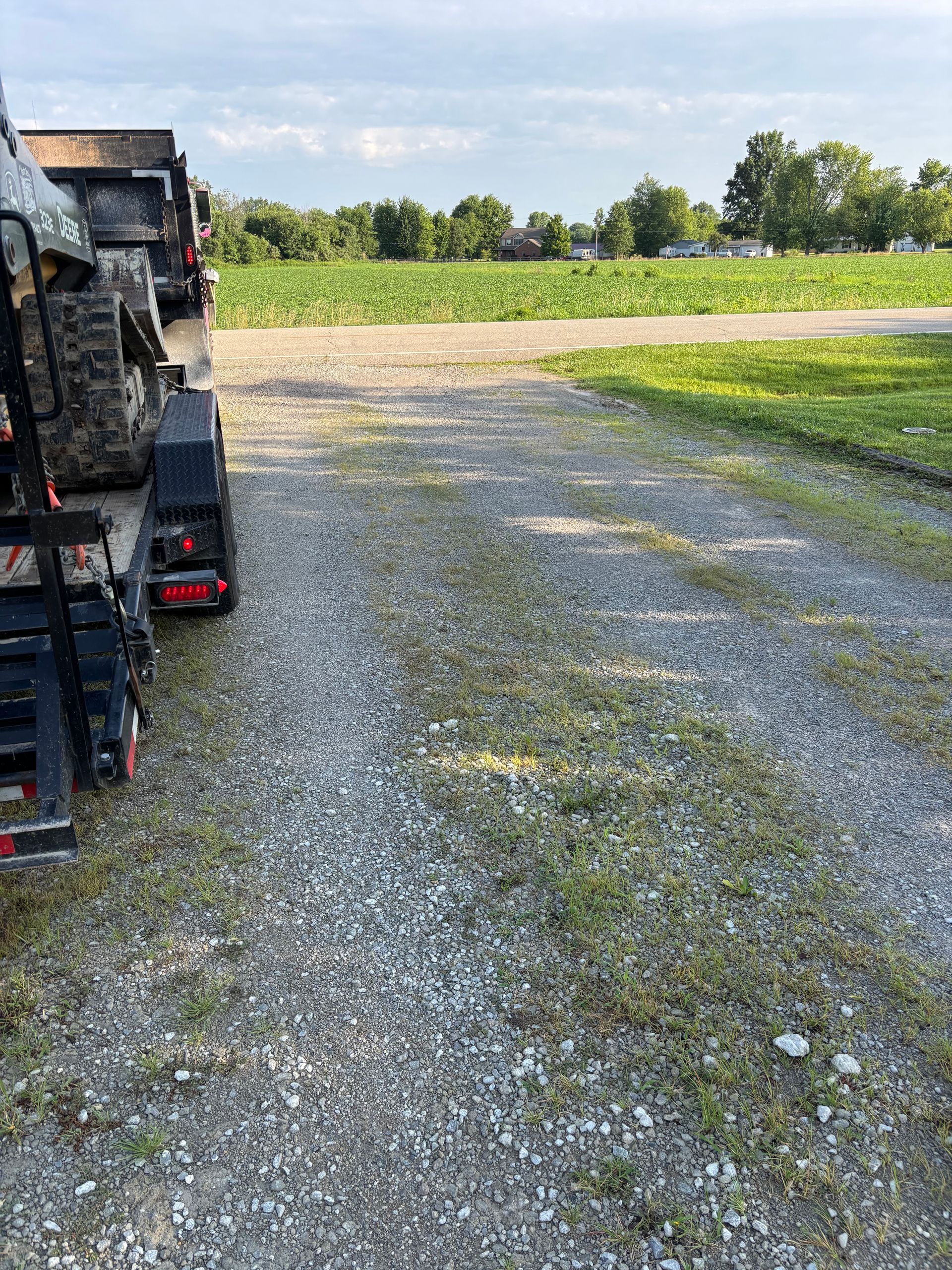 Gravel driveway beside a tractor trailer, opening onto a green field under a sunny sky.