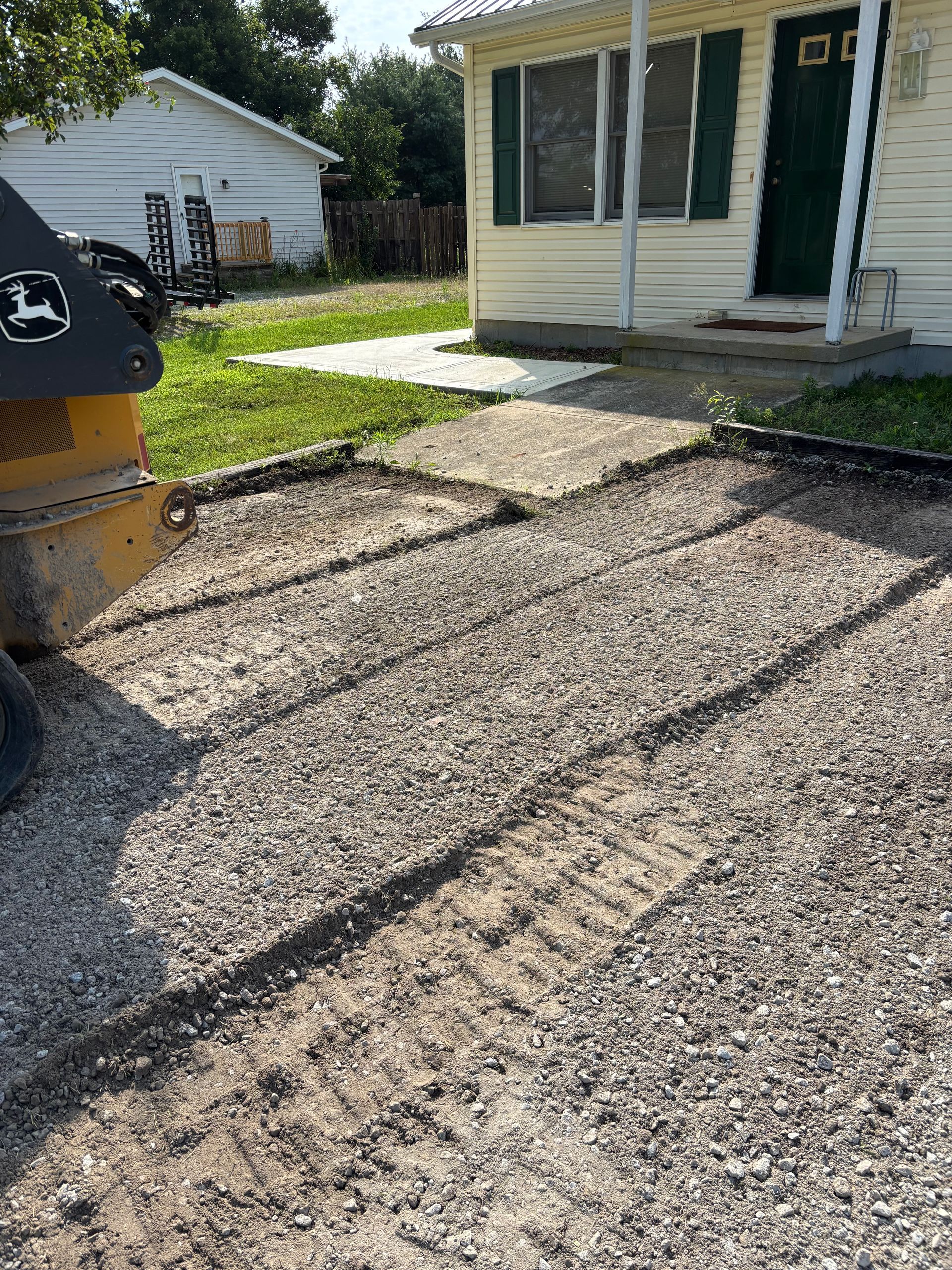 Driveway under repair beside a house, with a yellow construction machine and fresh asphalt patches.