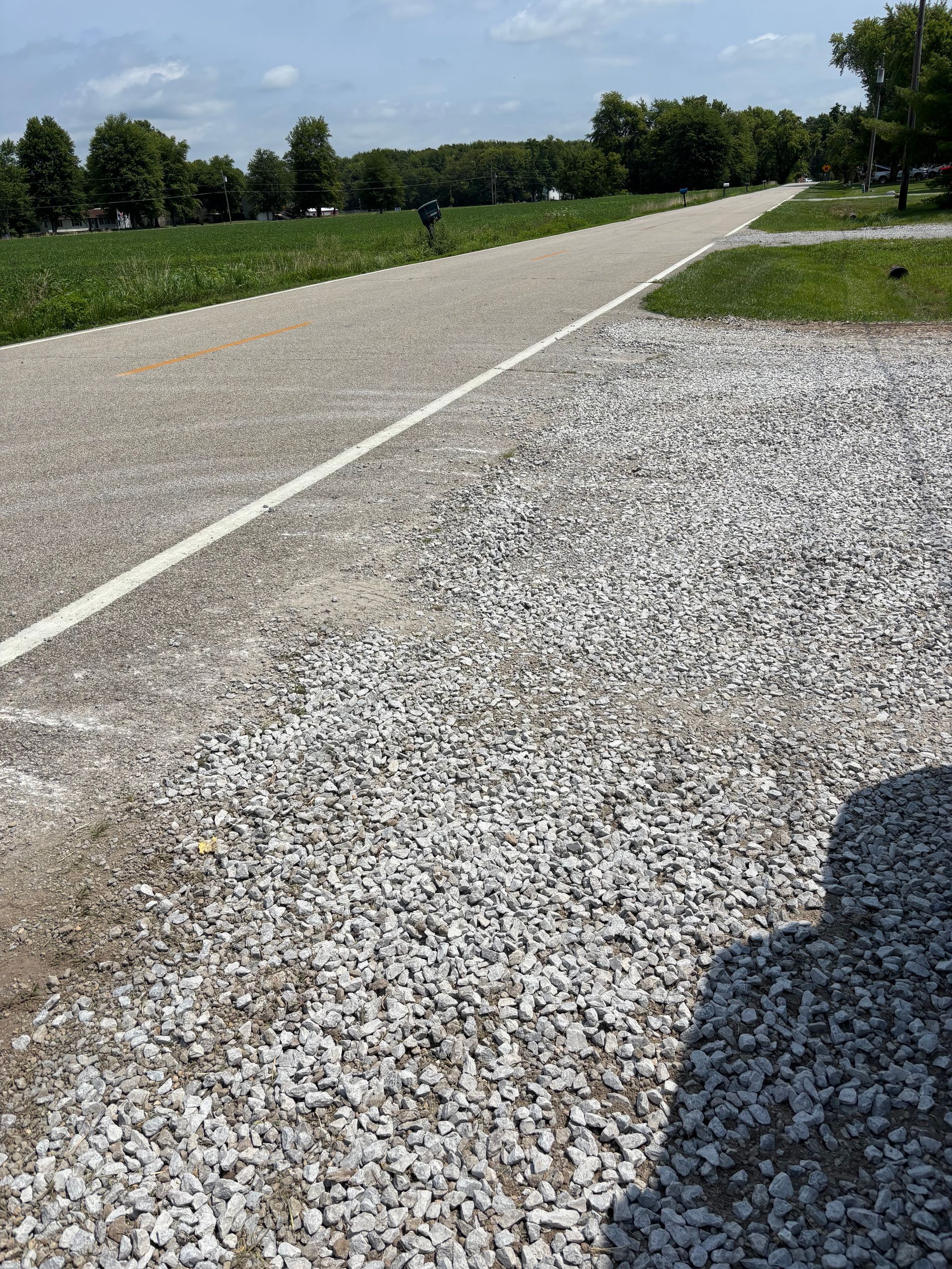 Gravel shoulder beside a quiet paved road with grassy fields and trees under a sunny sky