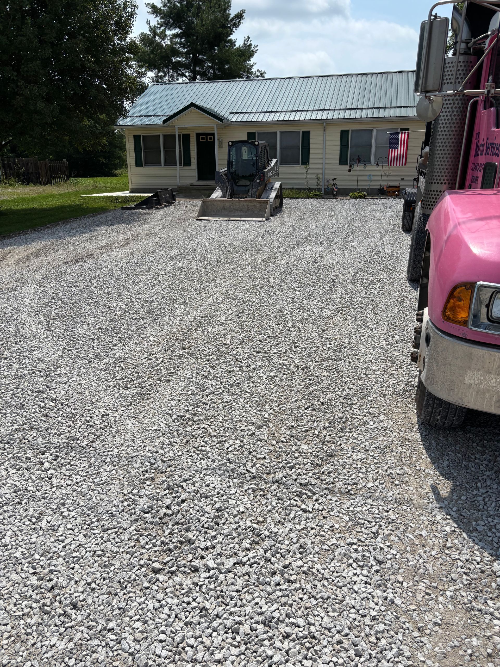 Gravel driveway leading to a small house, with a pink truck parked at right and a loader on the drive.