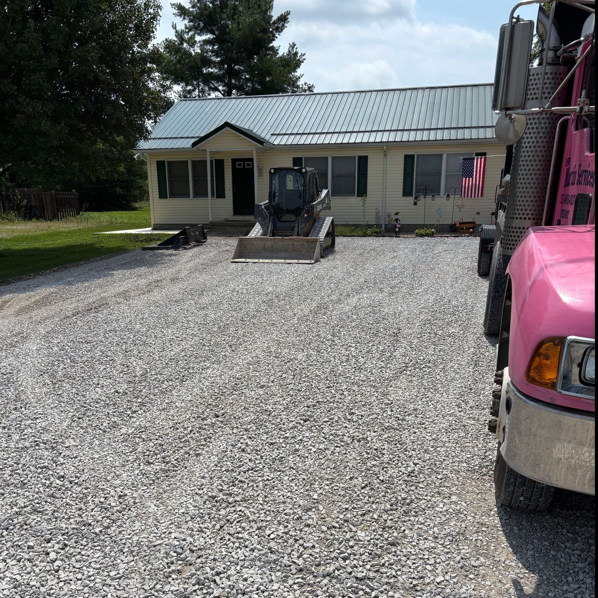 Gravel driveway leading to a beige house with a green roof and a pink truck in the foreground