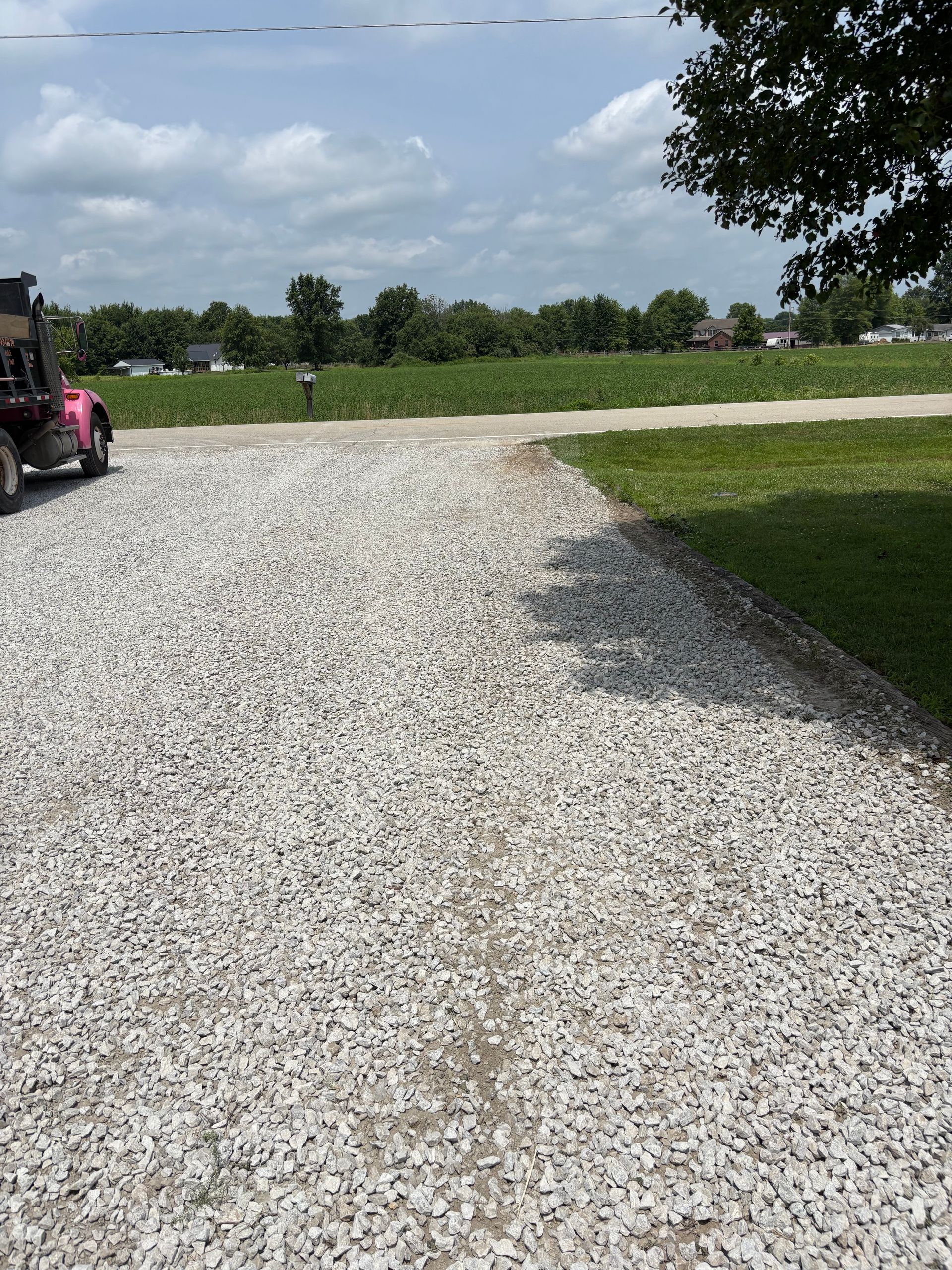 Gravel driveway curving through a grassy field under a cloudy sky, with a pink vehicle at left.