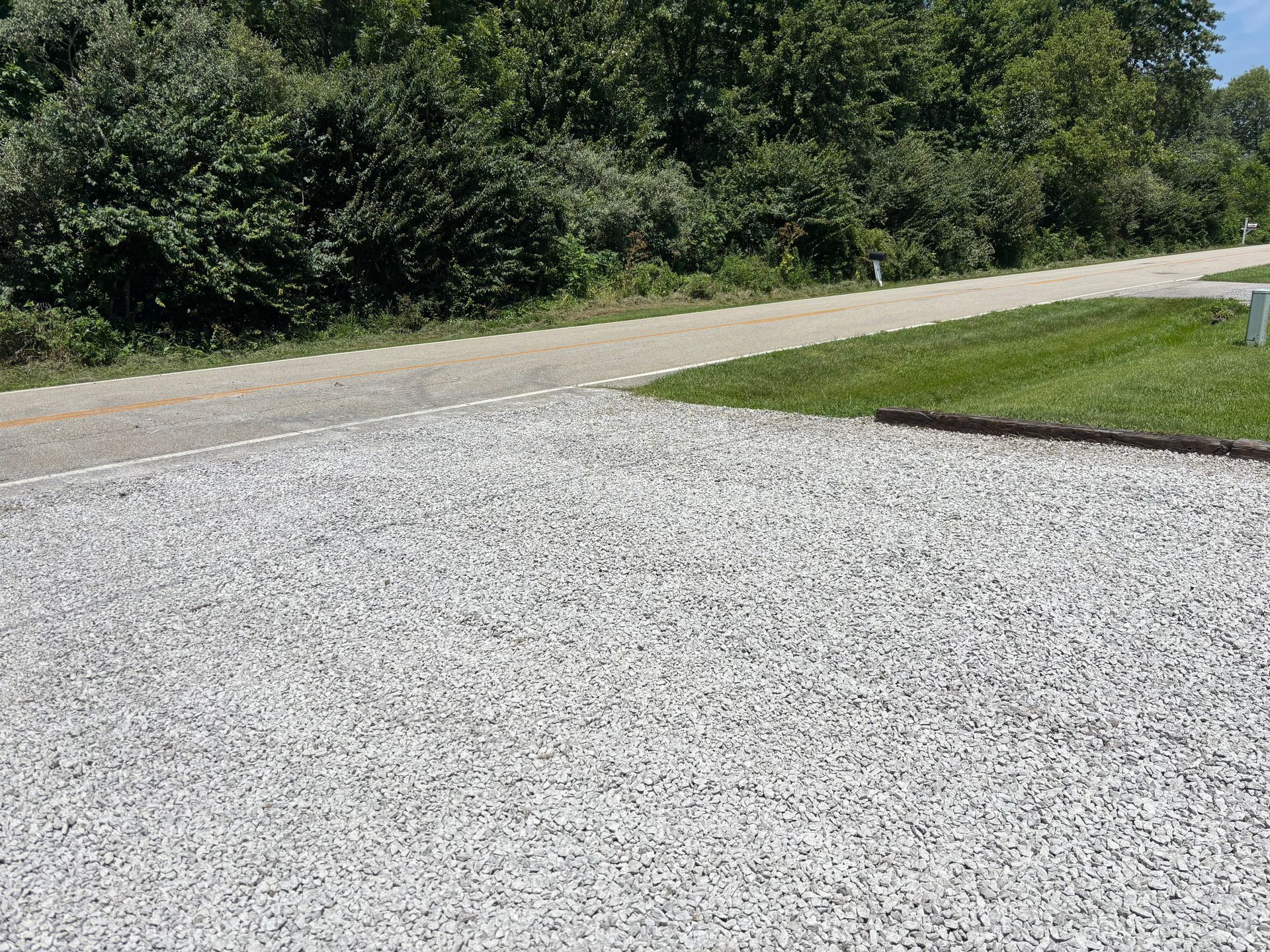 Gravel driveway beside a paved road, with green grass and trees in the background.
