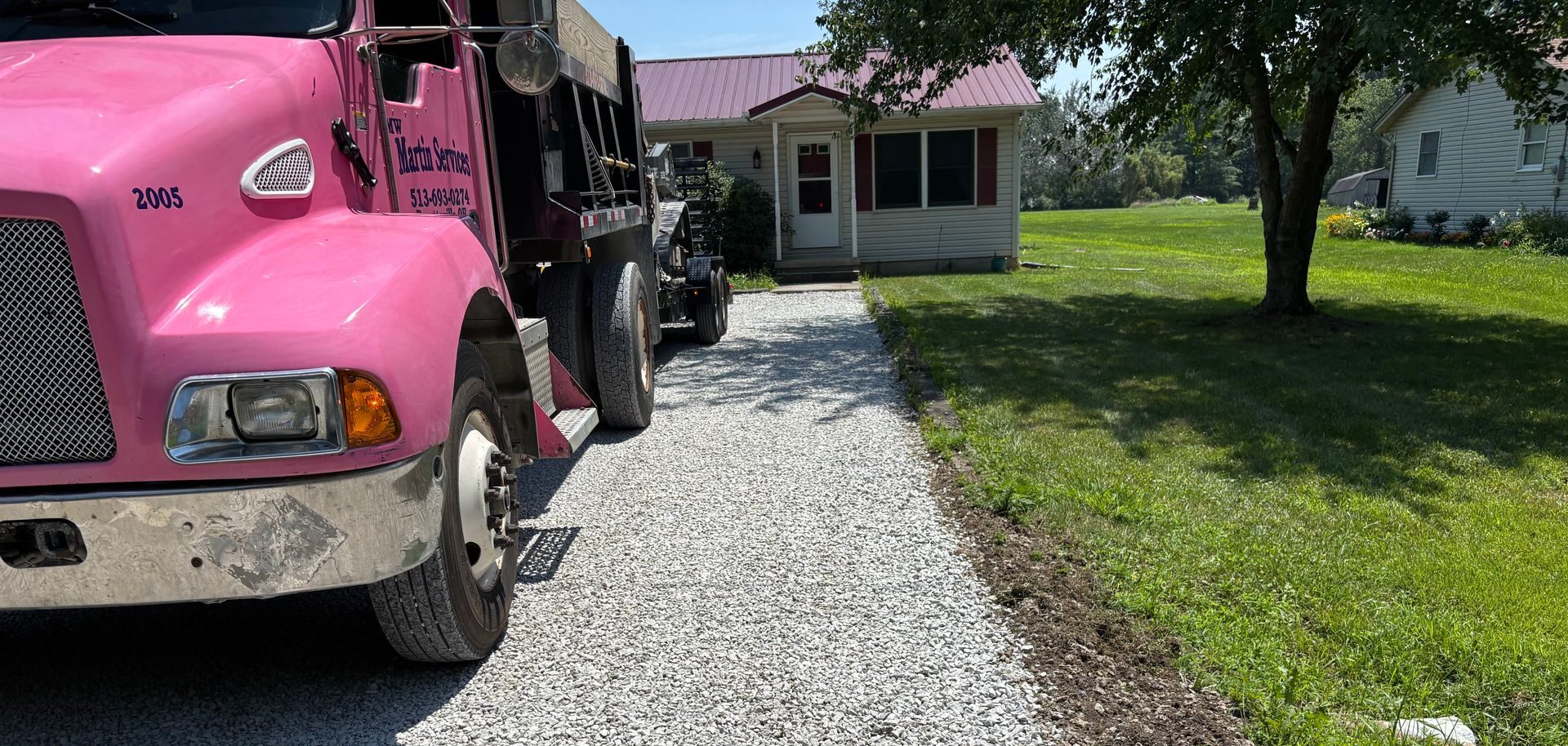 Pink dump truck on a gravel driveway beside a white house and green lawn