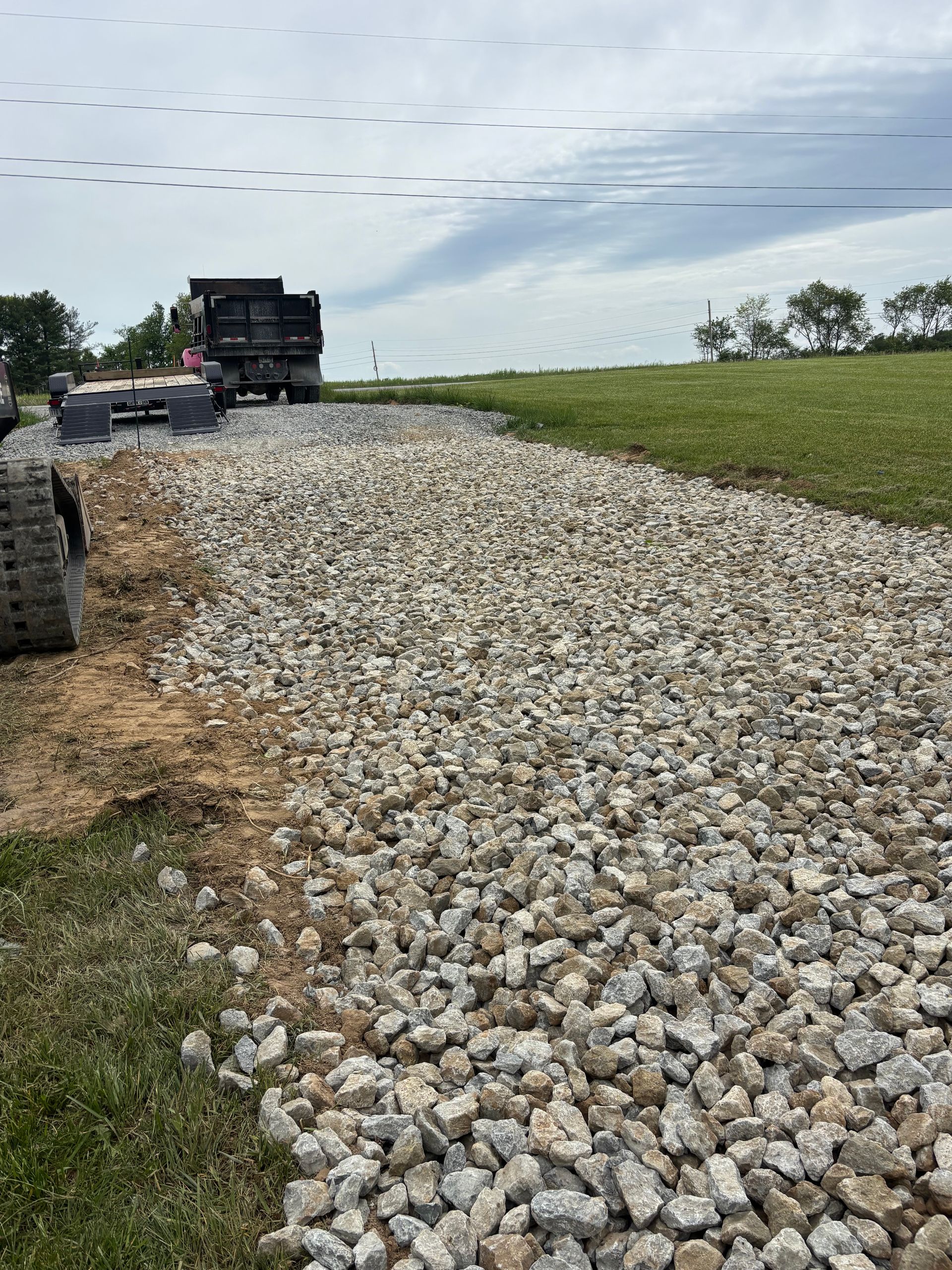 Gravel road under construction beside a grassy field, with heavy equipment and a dump truck in the distance.