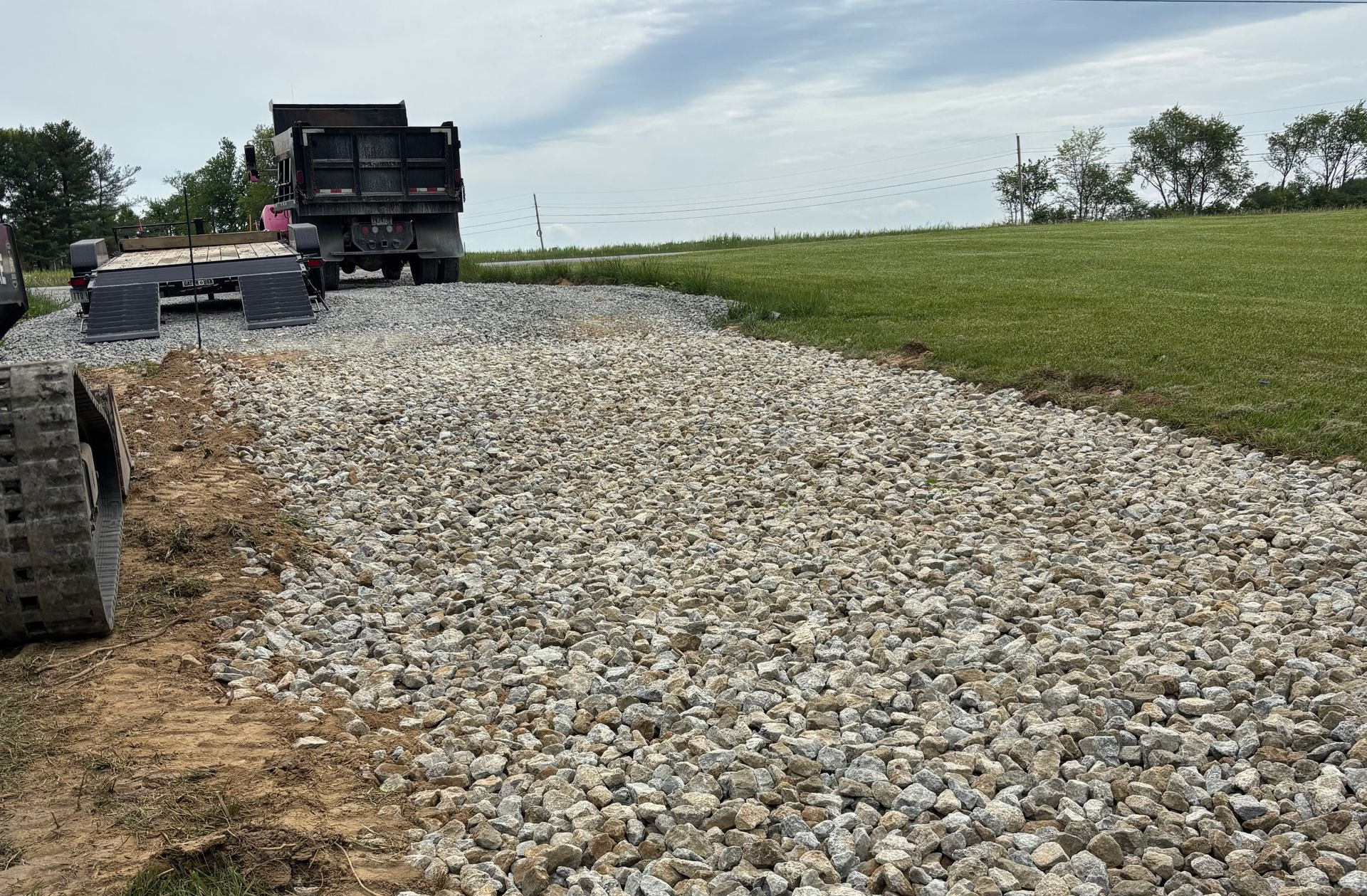Dump trucks on a gravel road beside a grassy field under a cloudy sky.