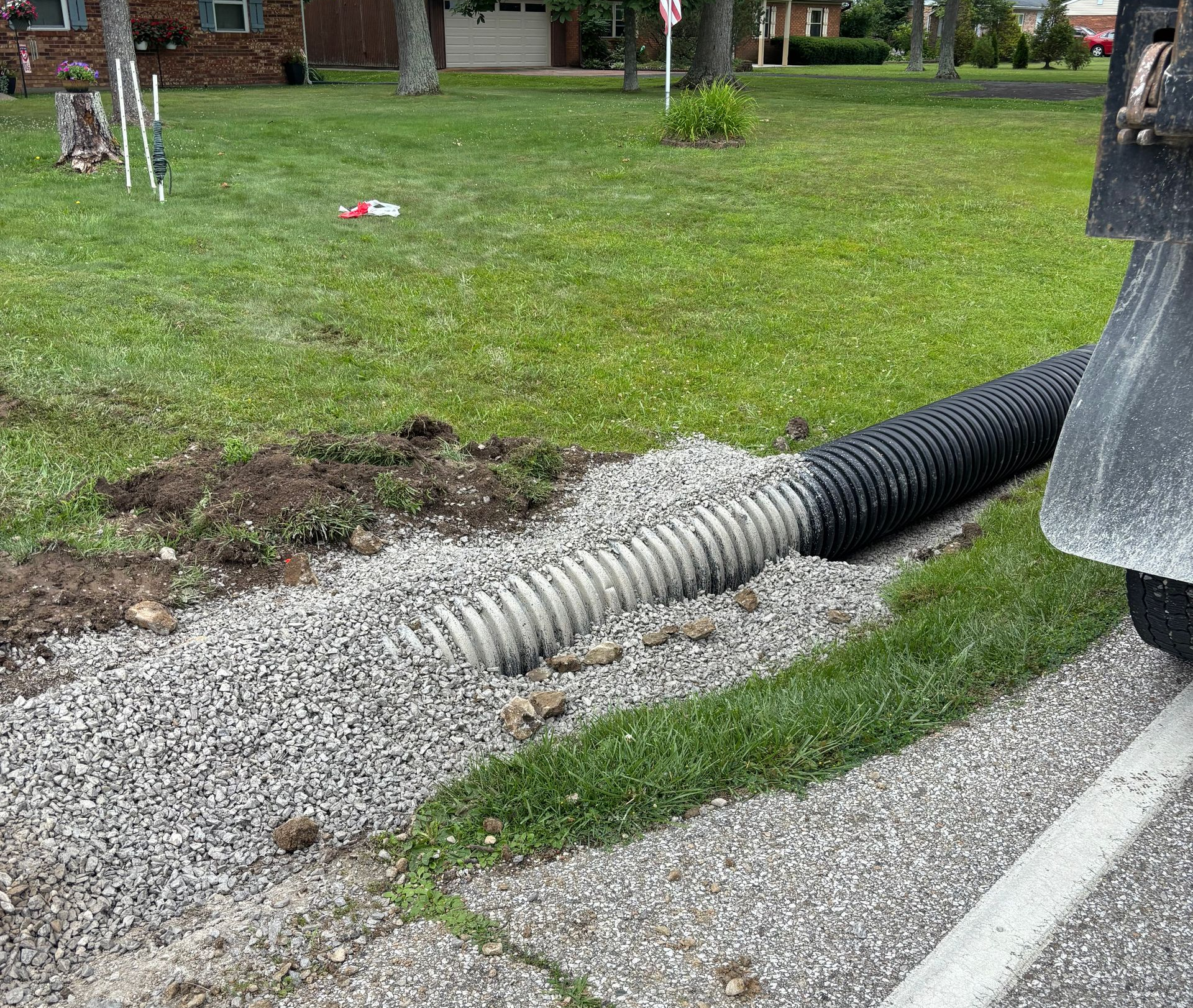 Black drainage pipe extending from curb into a grassy roadside ditch with gravel around it