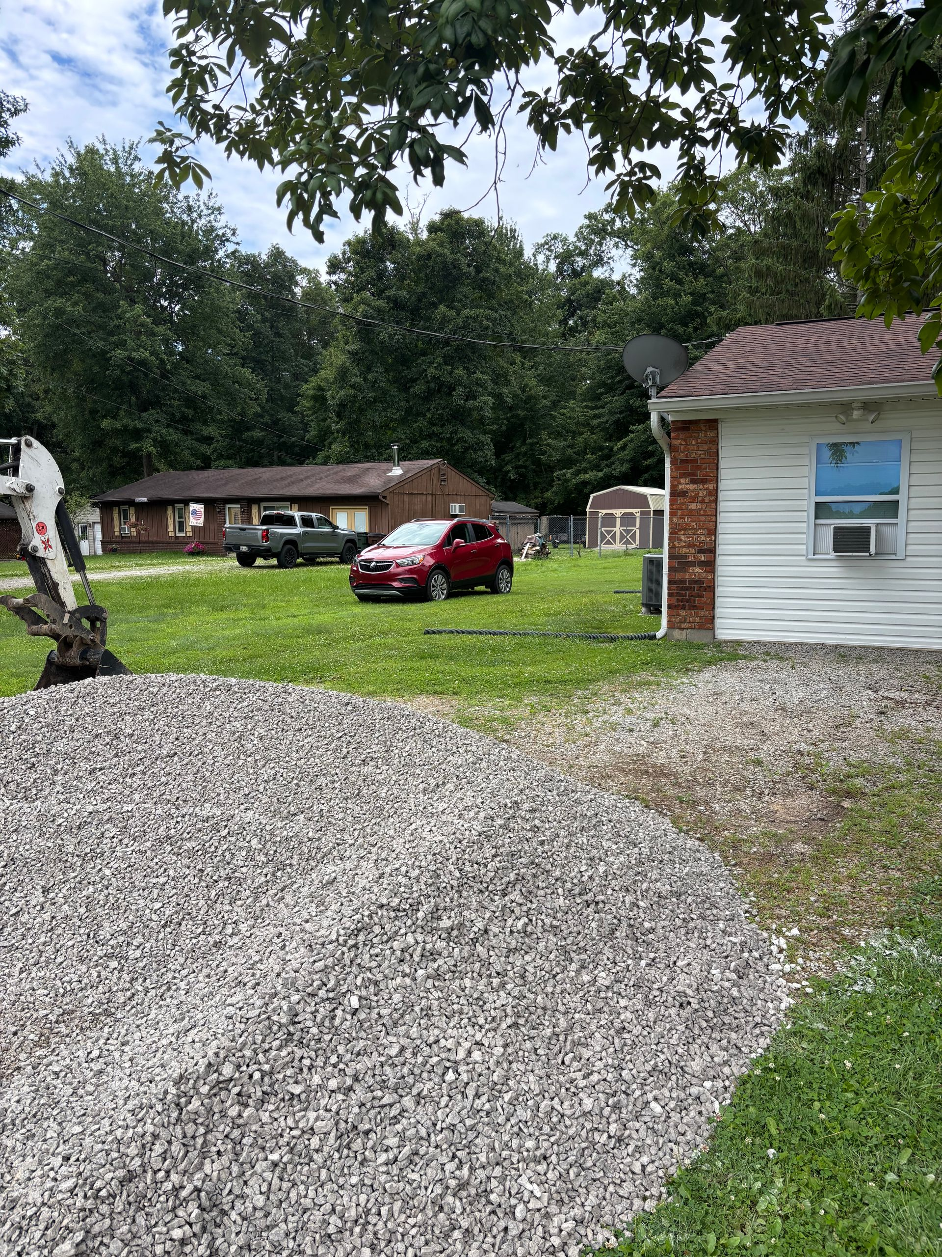 Gravel driveway beside a white outbuilding, with a red car parked on a grassy yard near a brick house.