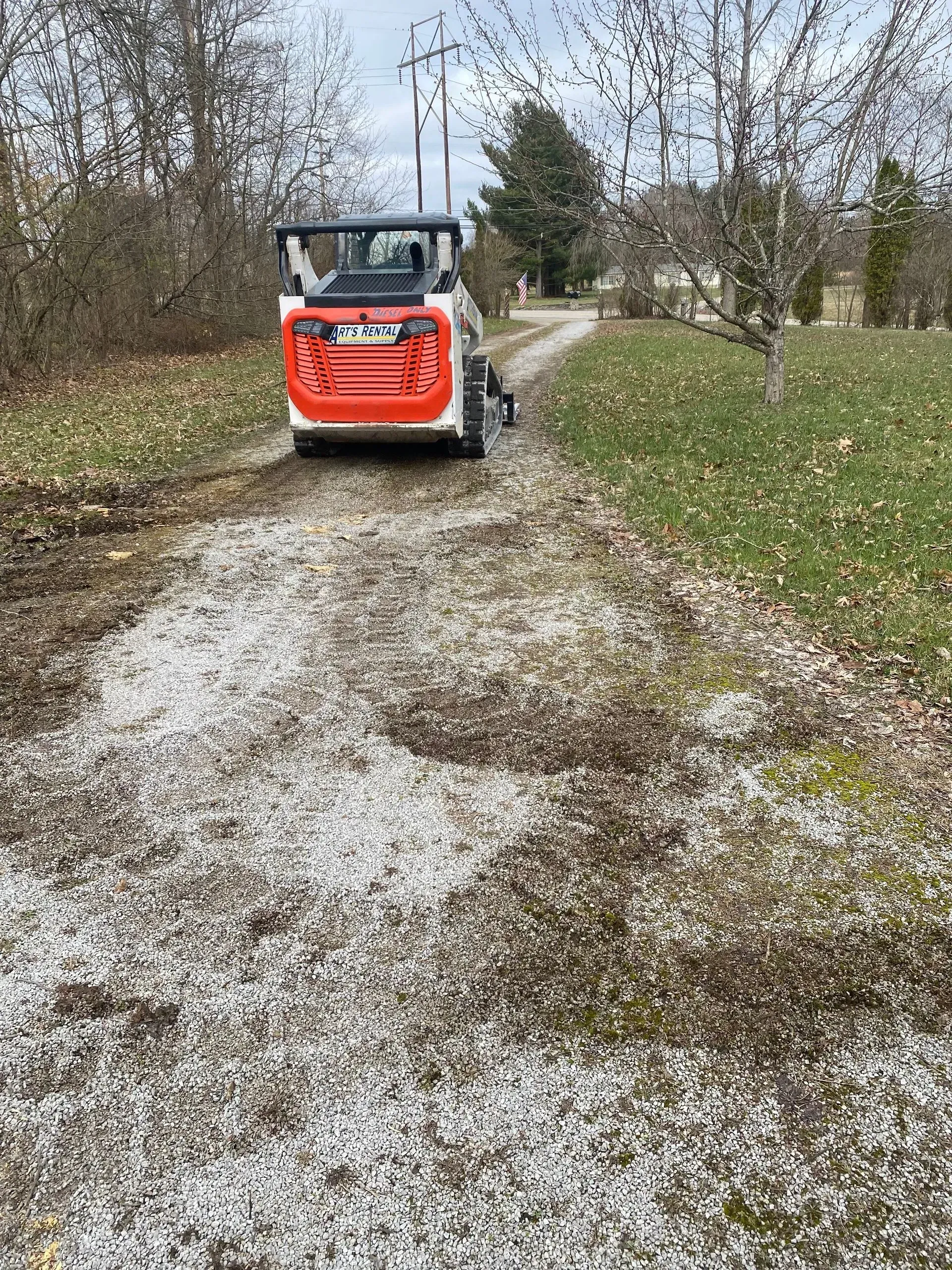 Red skid-steer loader on a gravel path beside grass and trees on a cloudy day