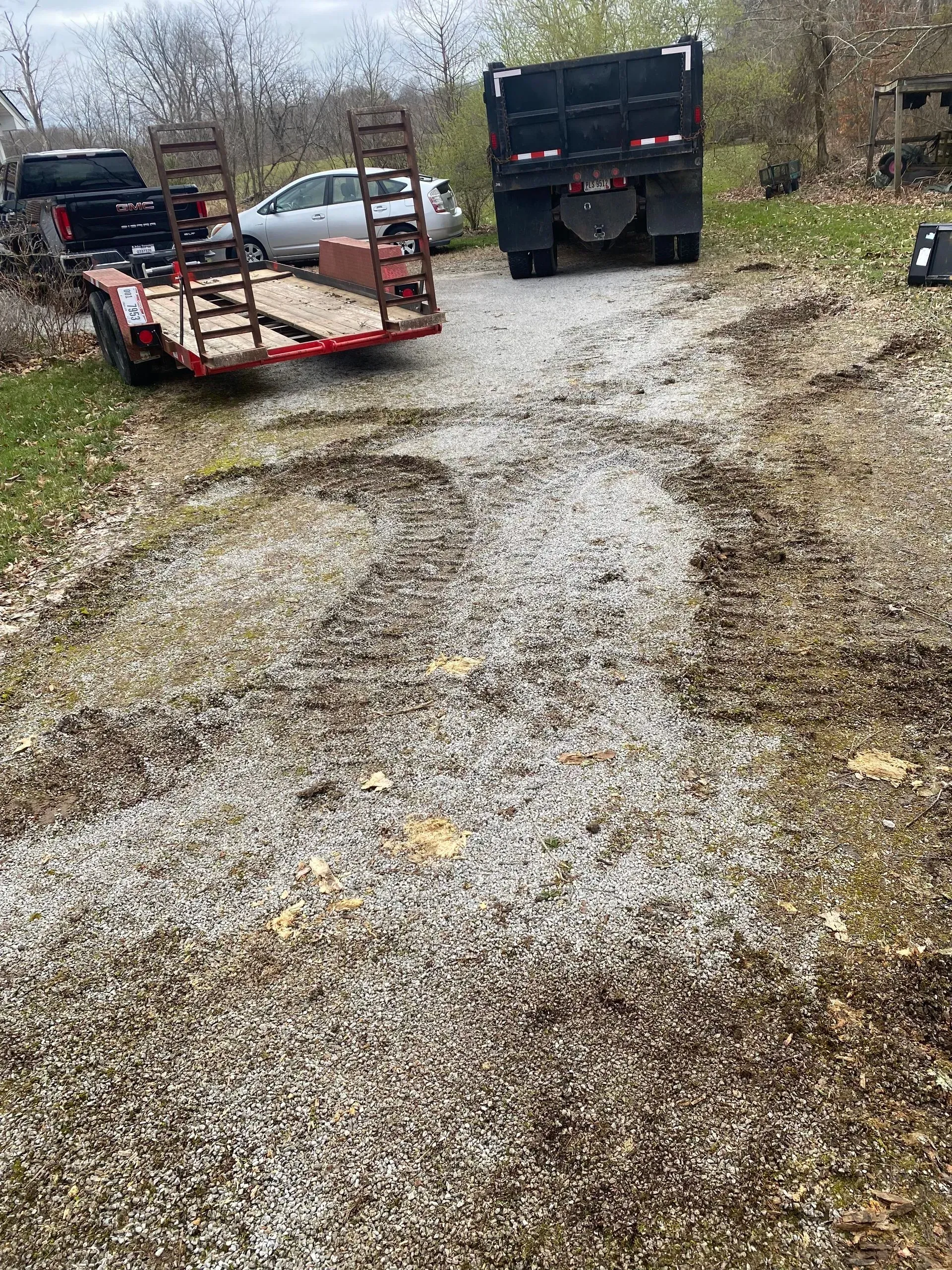 Gravel driveway with large muddy ruts, a flatbed trailer, and a dump truck under overcast skies