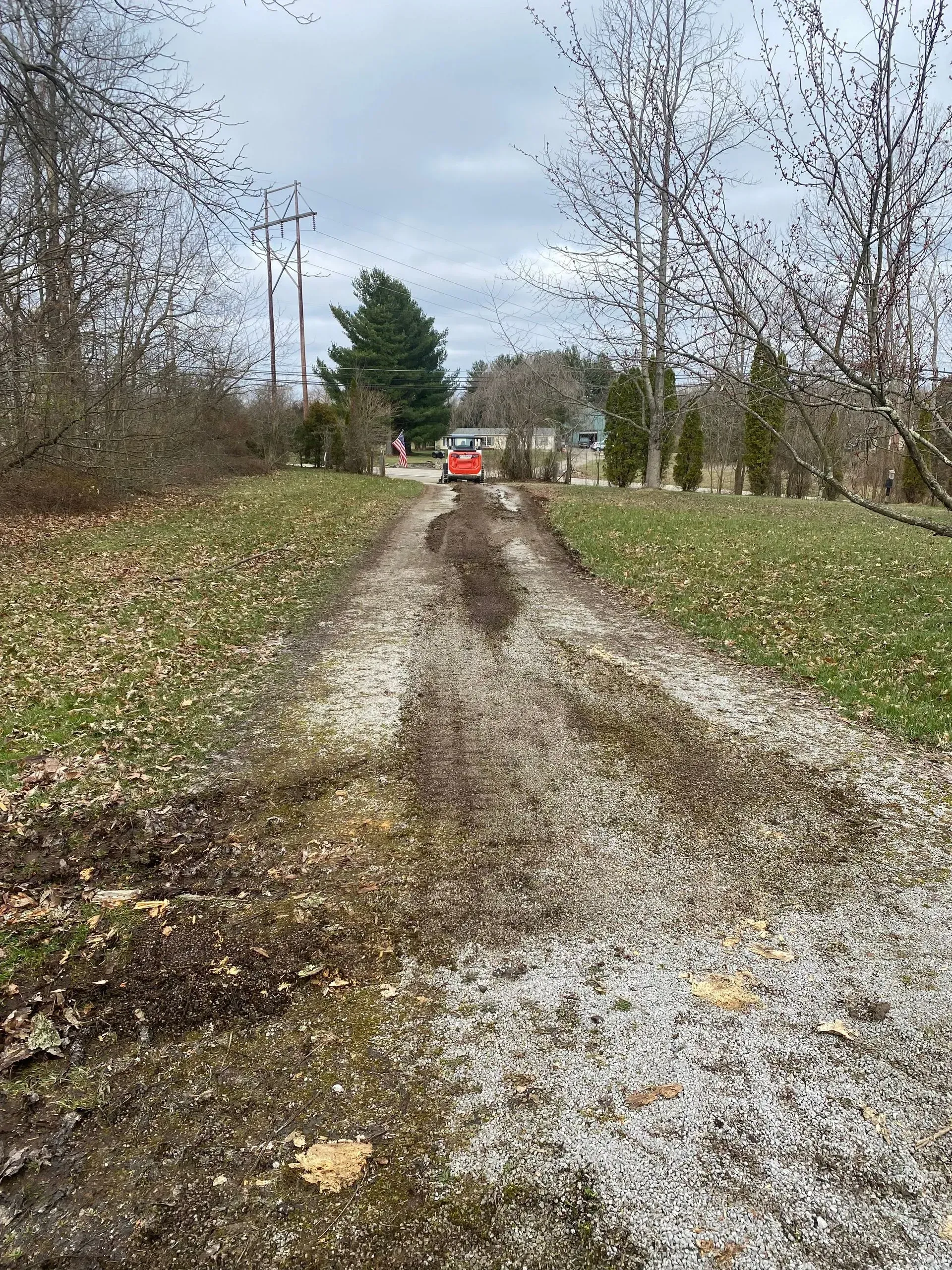 Muddy gravel driveway lined with bare trees, leading to a red tractor in the distance