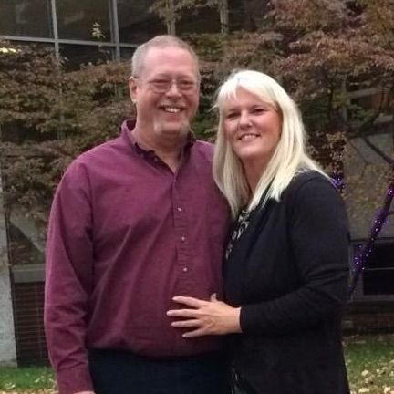Smiling couple posing outdoors in front of a building and trees, wearing maroon and black tops