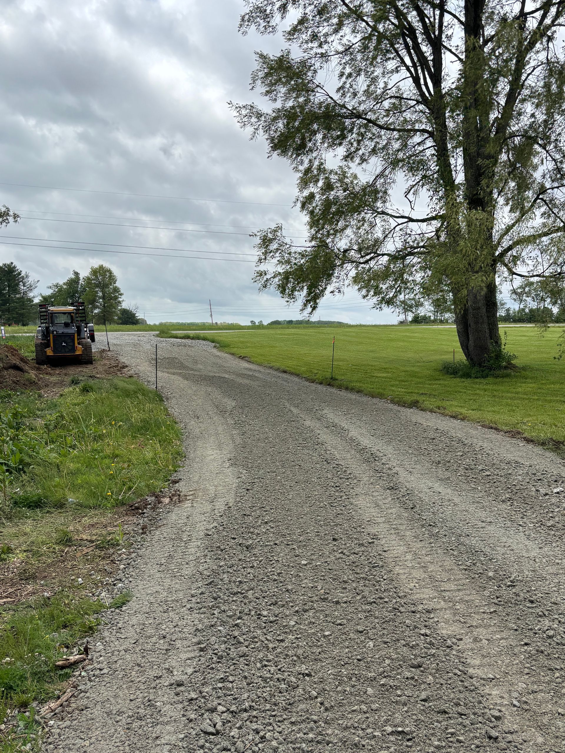 Gravel road curving through a green field under cloudy sky, with a tractor at the left edge.