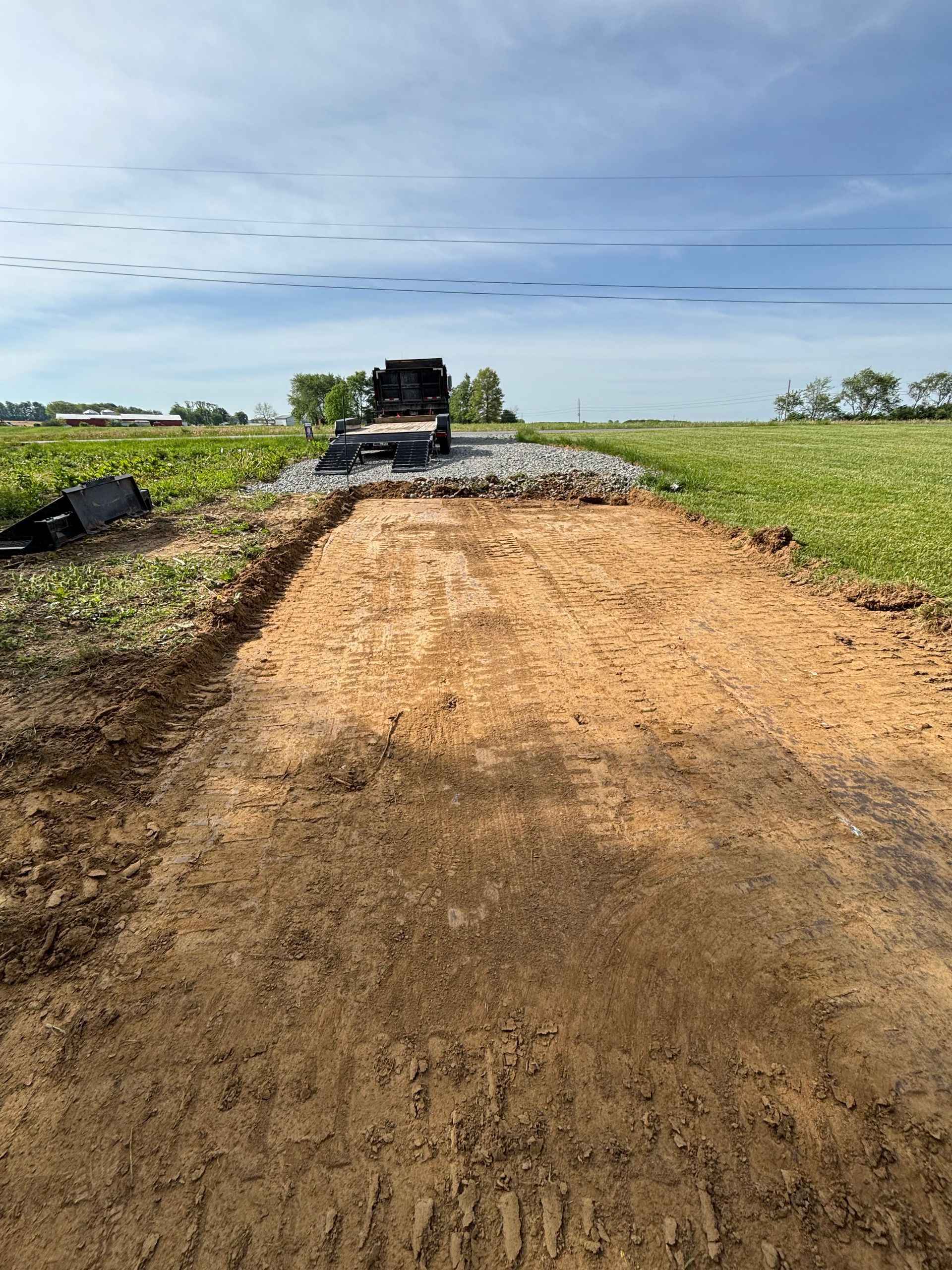 A dirt road under construction in a flat rural field, with a dump truck at the far end and blue sky overhead