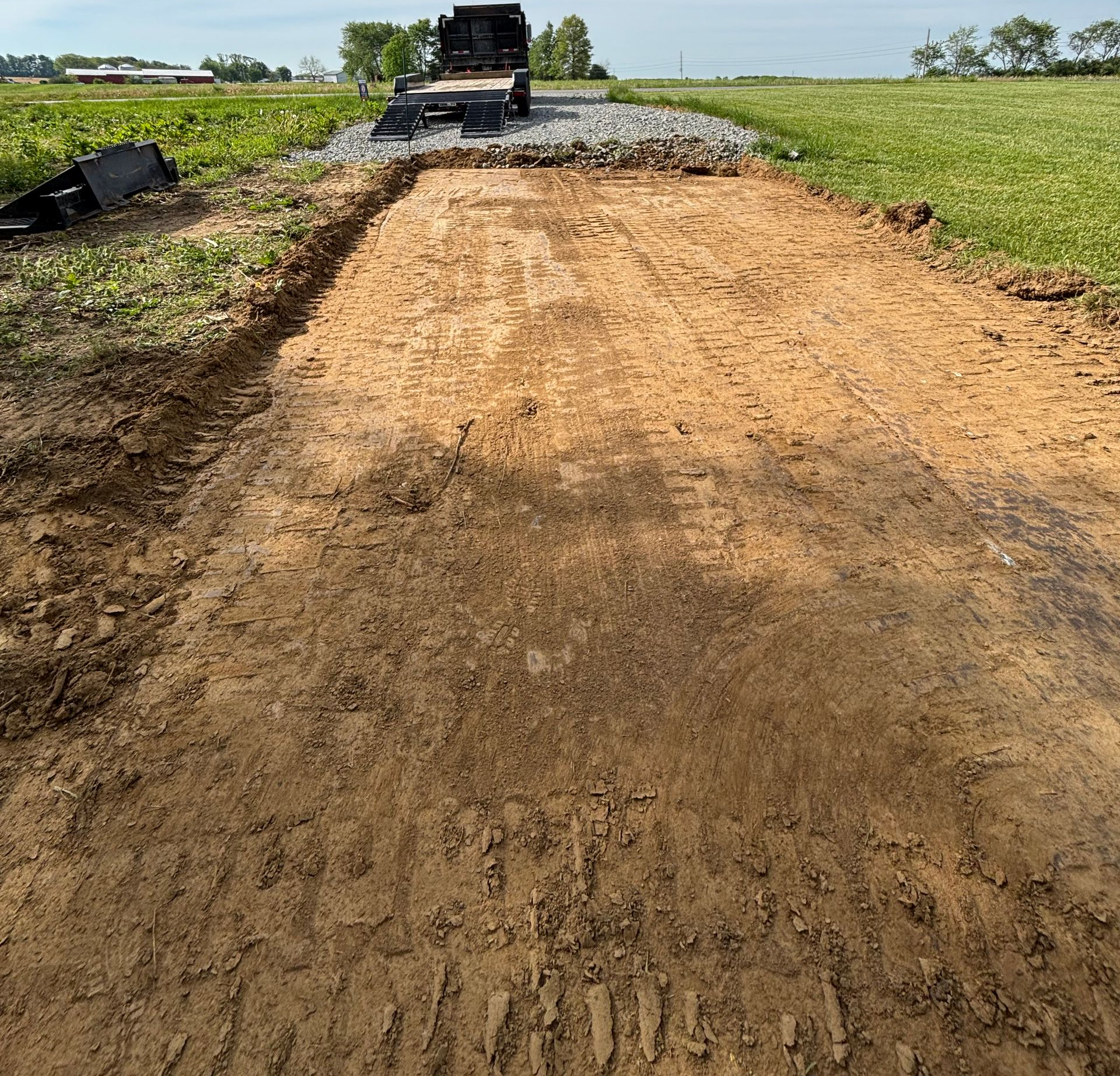 Graded dirt road under construction with a dump truck and gravel pile ahead in a rural field
