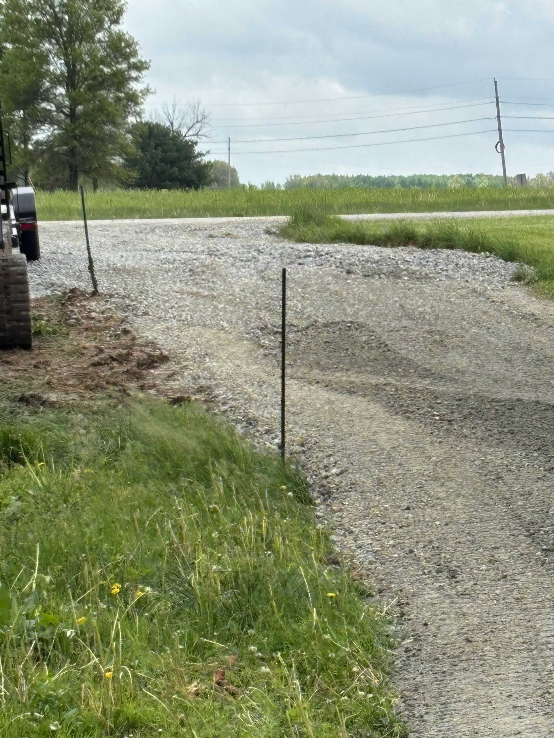 Gravel road curving past a grassy field with trees and utility poles on an overcast day
