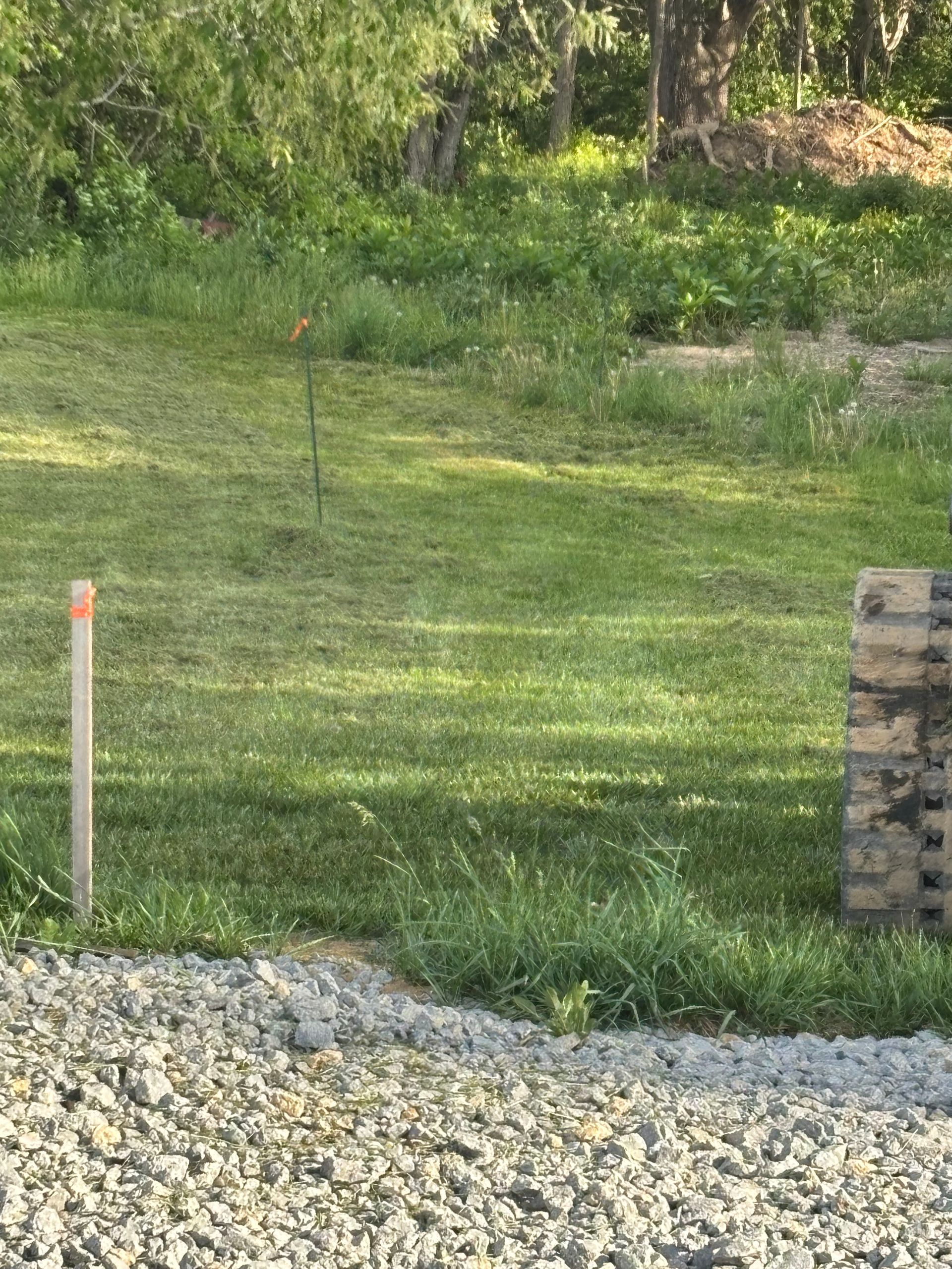 Grassy yard with a gravel driveway, a utility stake, and a stacked stone pillar beside trees.
