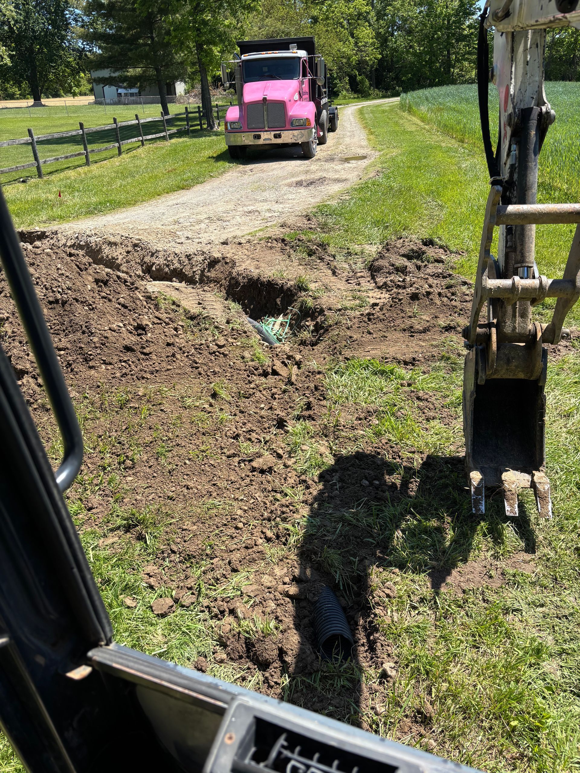 Pink truck on a dirt road by a grassy ditch, seen from inside a construction vehicle with an excavator arm visible
