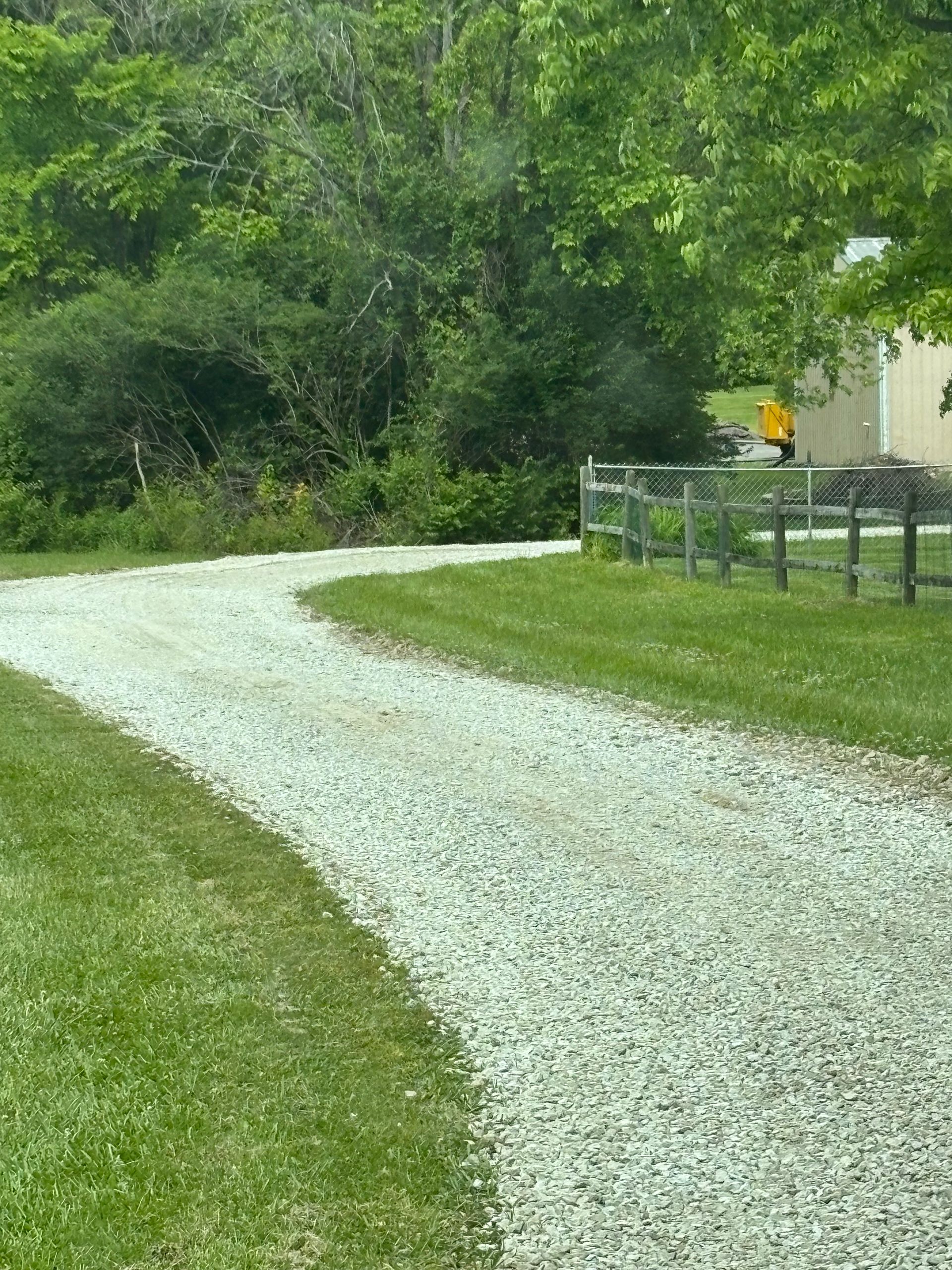 Gravel driveway curving through green grass and trees beside a white building and fence